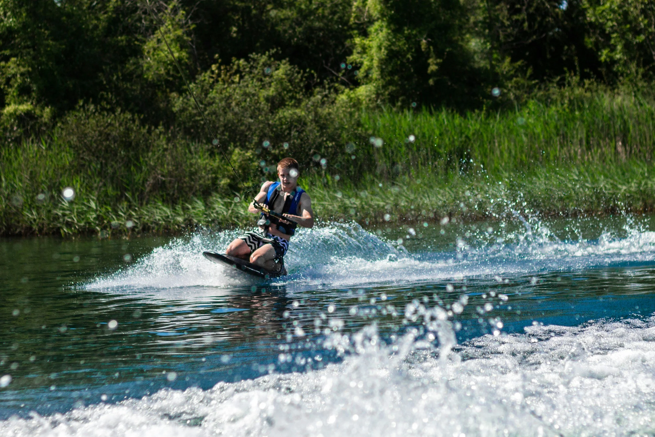 A boy wearing a life jacket and striped swim trunks is wakeboarding on a river surrounded by green trees.