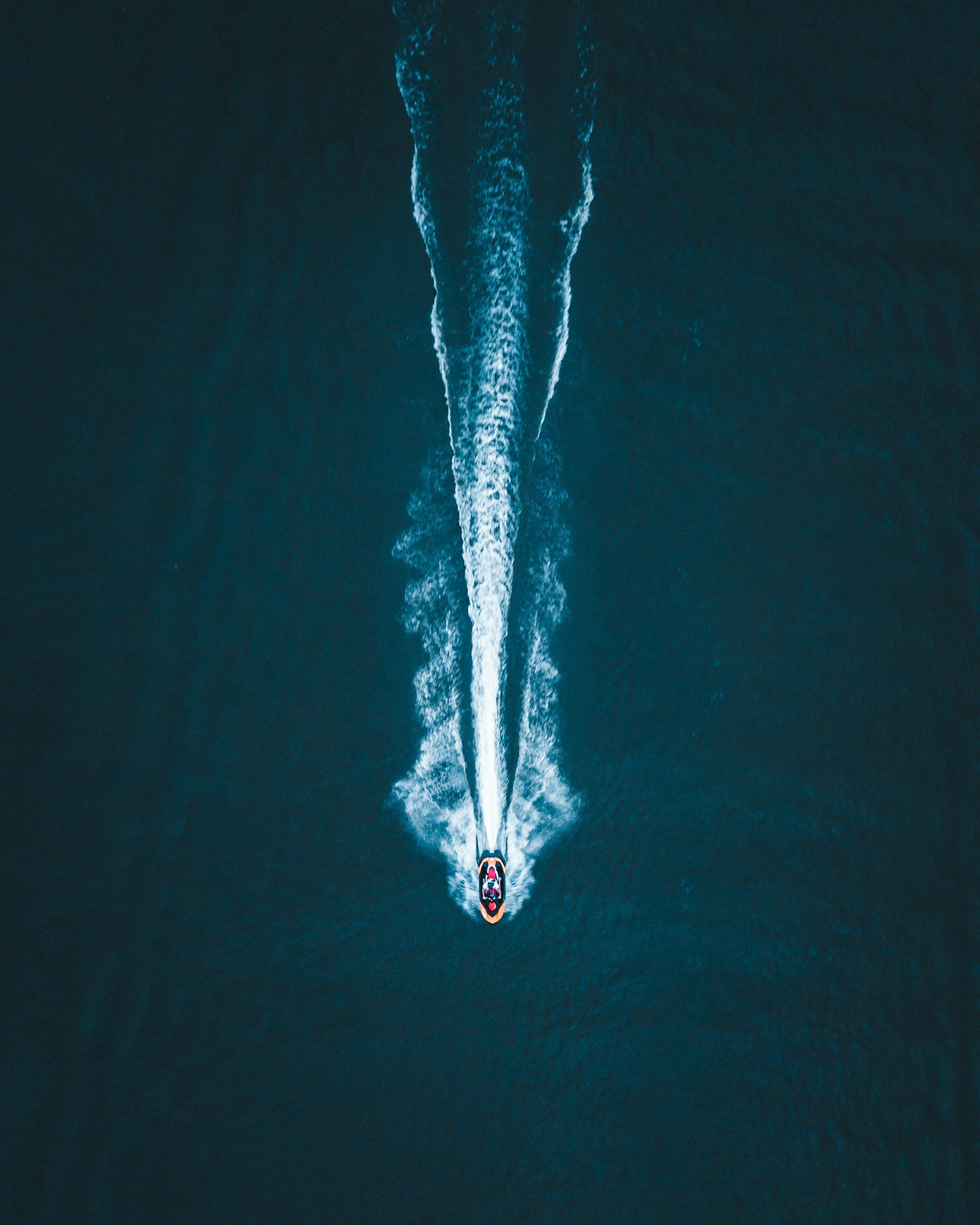An aerial view of a speedboat creating a long white wake as it moves across the dark blue water.