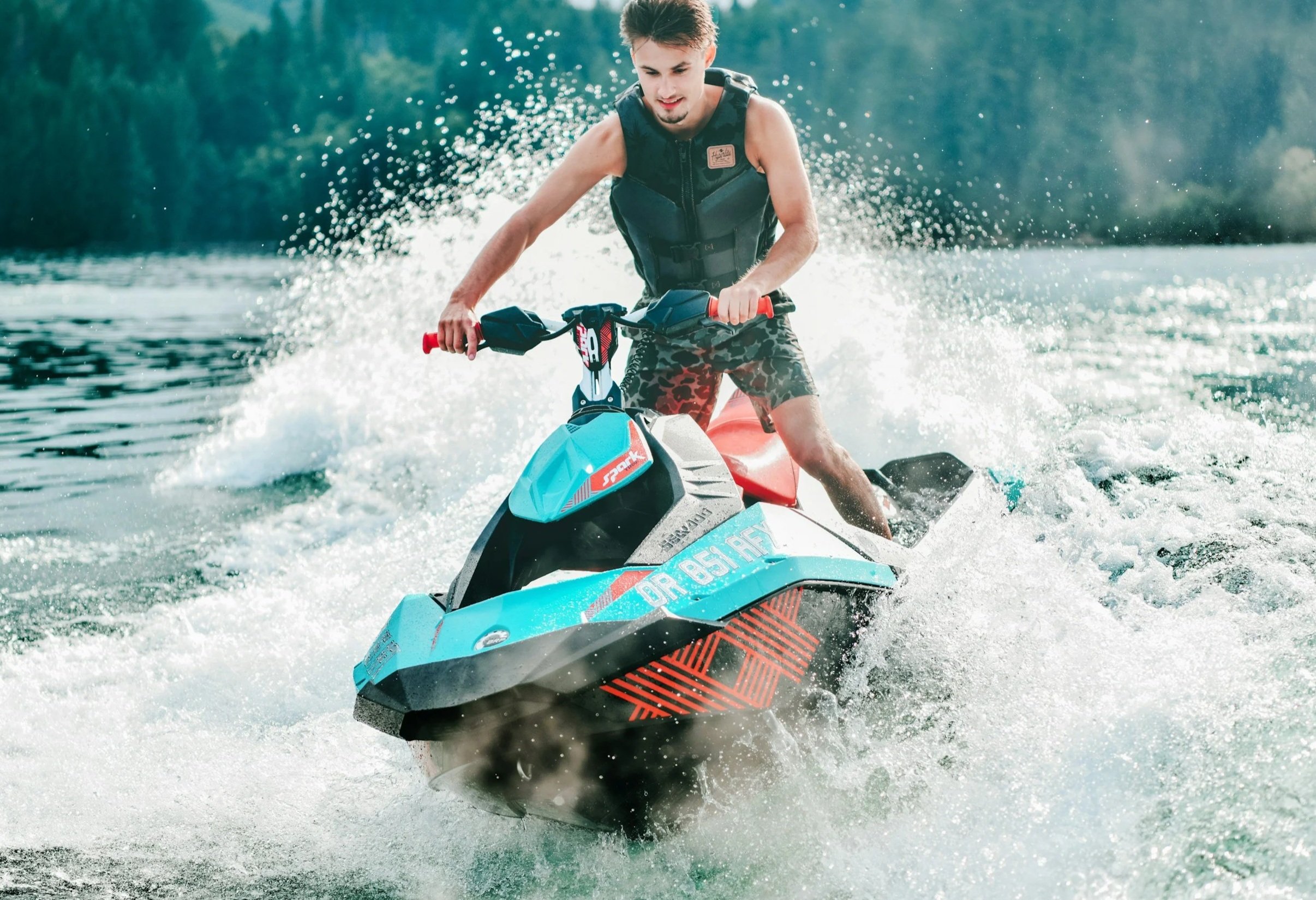 Young man riding a blue and black jet ski on a lake, wearing a life jacket and camo shorts, with water splashing behind him.
