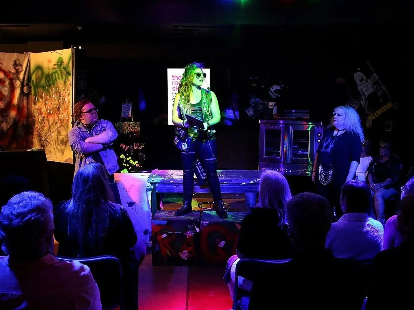 A theatrical performance on a small stage with a woman in punk fashion holding a book, standing on a table illuminated by colorful stage lights, with three other women watching and audience members seated in front.