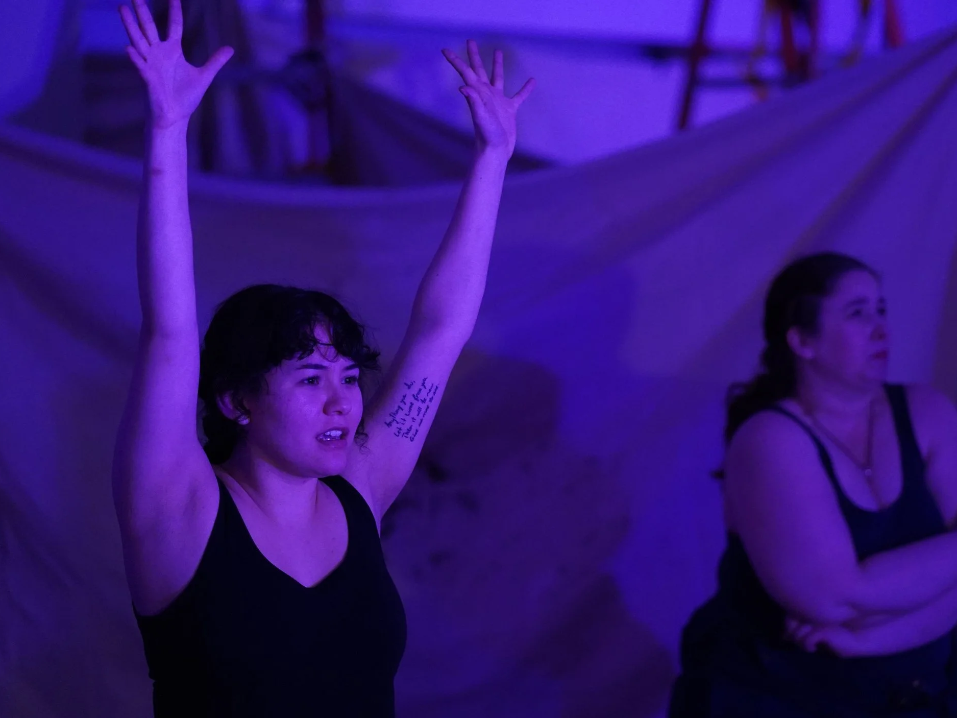A young woman with short dark hair and a tattoo on her arm raising both hands with open palms in a dimly lit room with purple lighting, and another woman standing in the background.