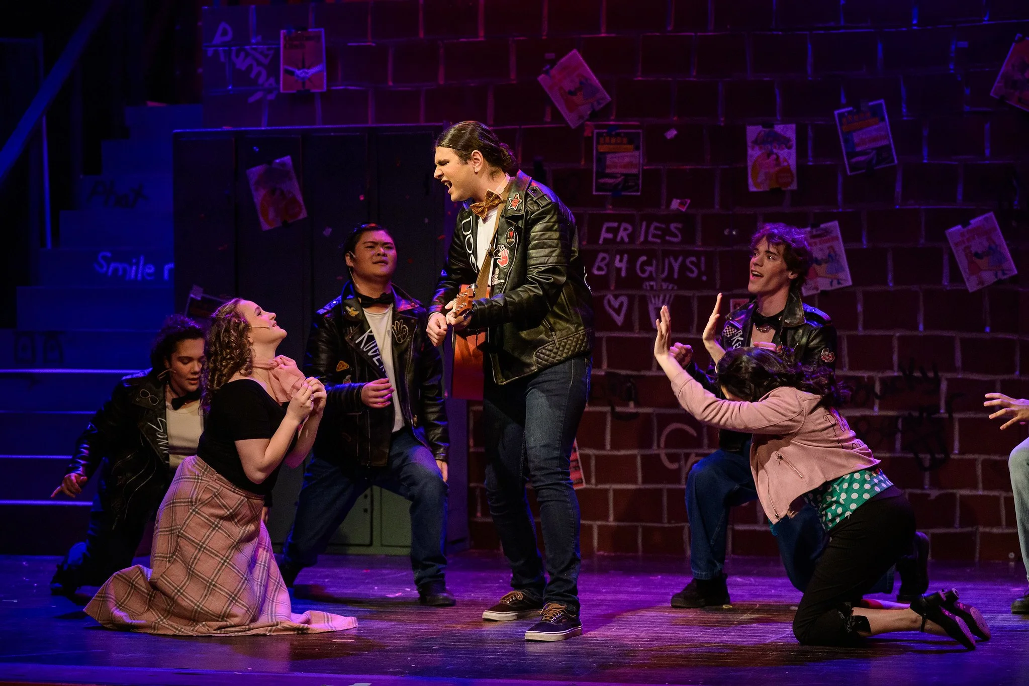 The scene is from a theatrical performance with six actors on stage. One actor, standing and playing a guitar, is engaging with the others, who are kneeling or sitting around her. The background shows a brick wall decorated with colorful posters and 