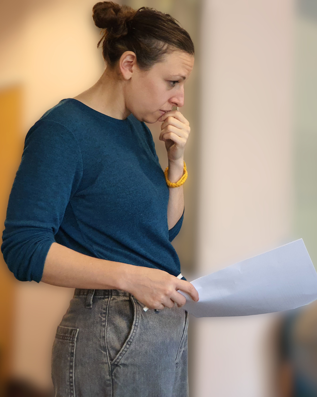 A woman with brown hair tied in a bun looking at a piece of paper with a worried expression, wearing a dark blue long-sleeve shirt and gray jeans.