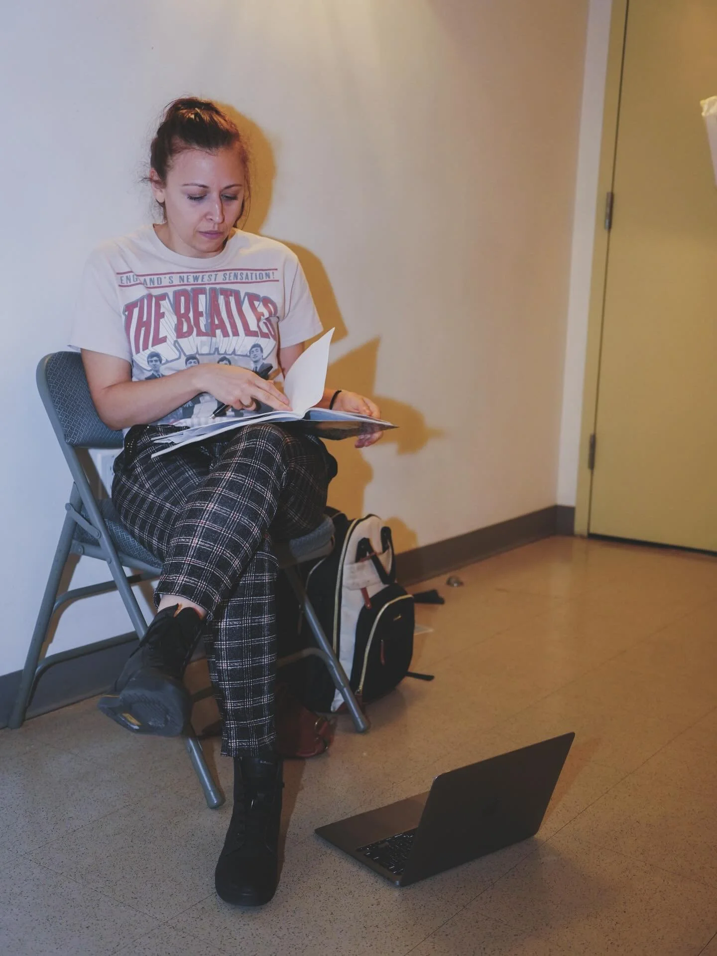 A woman sitting on a gray folding chair, reading a booklet with a relaxed posture. She is wearing a Beatles t-shirt, plaid pants, and black shoes, with a backpack and laptop on the floor nearby in a room with plain walls and a closed yellow door.