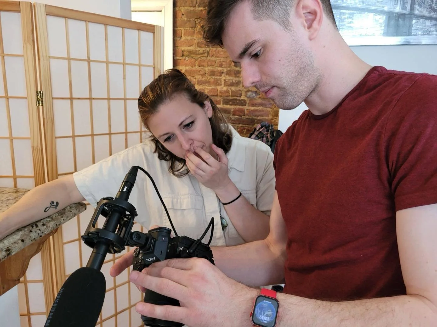 Two individuals, a woman and a man, closely examining a camera together indoors. The woman has her hand over her mouth in surprise or concern. The man is holding some part of the camera and is wearing a smartwatch. The background features a privacy screen, a brick wall, and a window.
