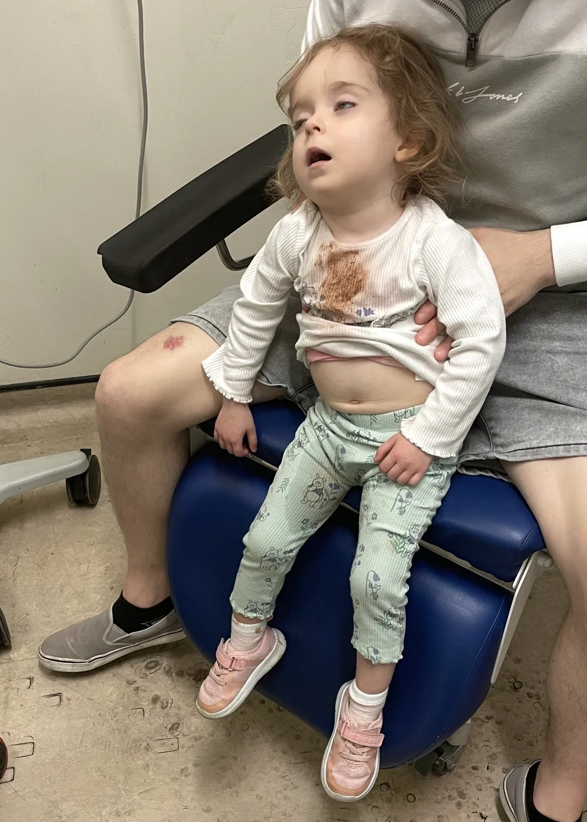 Young girl sitting on a chair with a tired or unwell expression, with a brown stain on her shirt, and scrape on her knee. An adult holds her gently, supporting her.