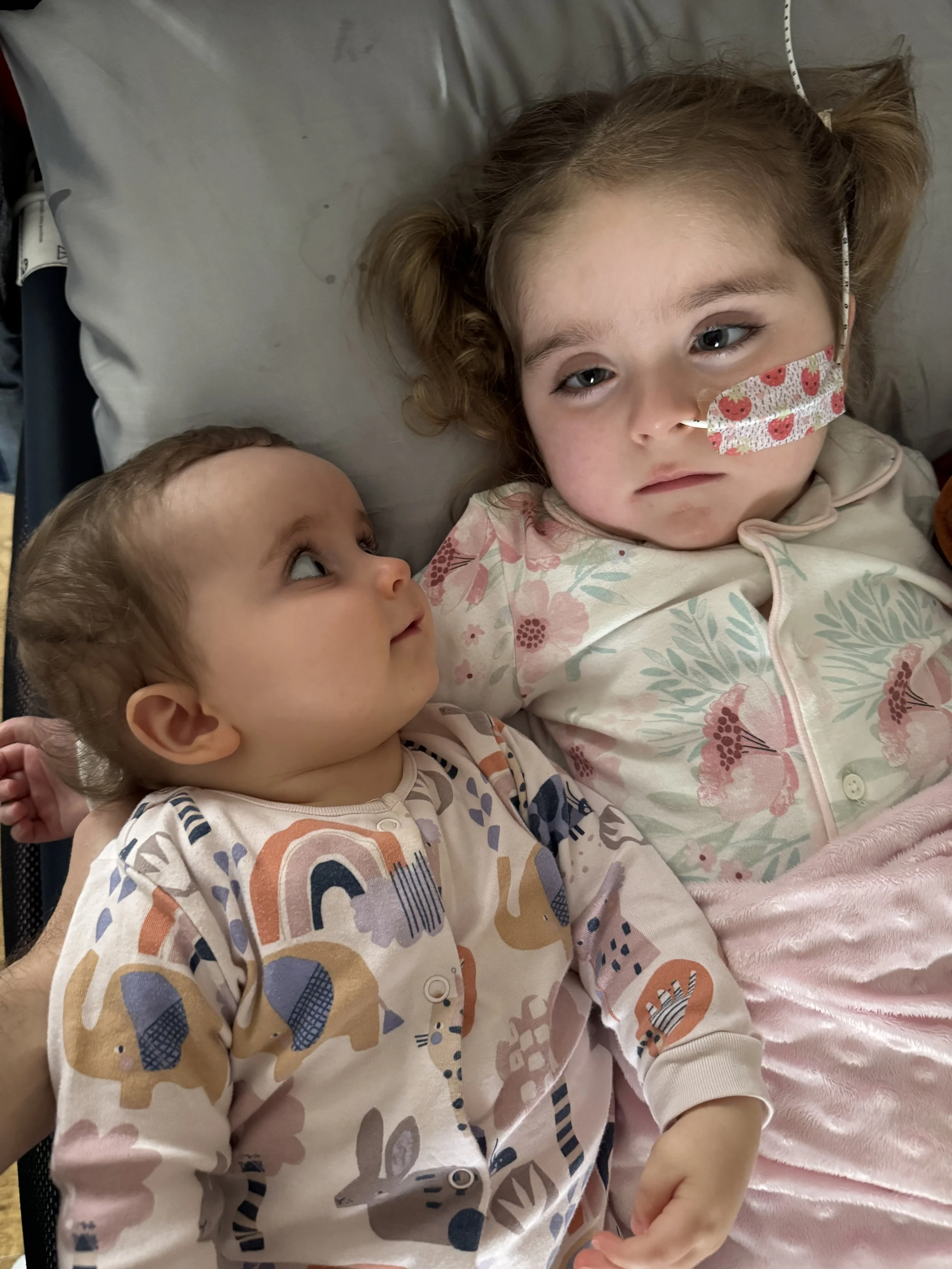 Two young children lying in hospital beds, with the girl wearing a floral hospital gown, a nasal oxygen tube, and a patterned face mask, while the boy wears a colorful top with animal and rainbow patterns.