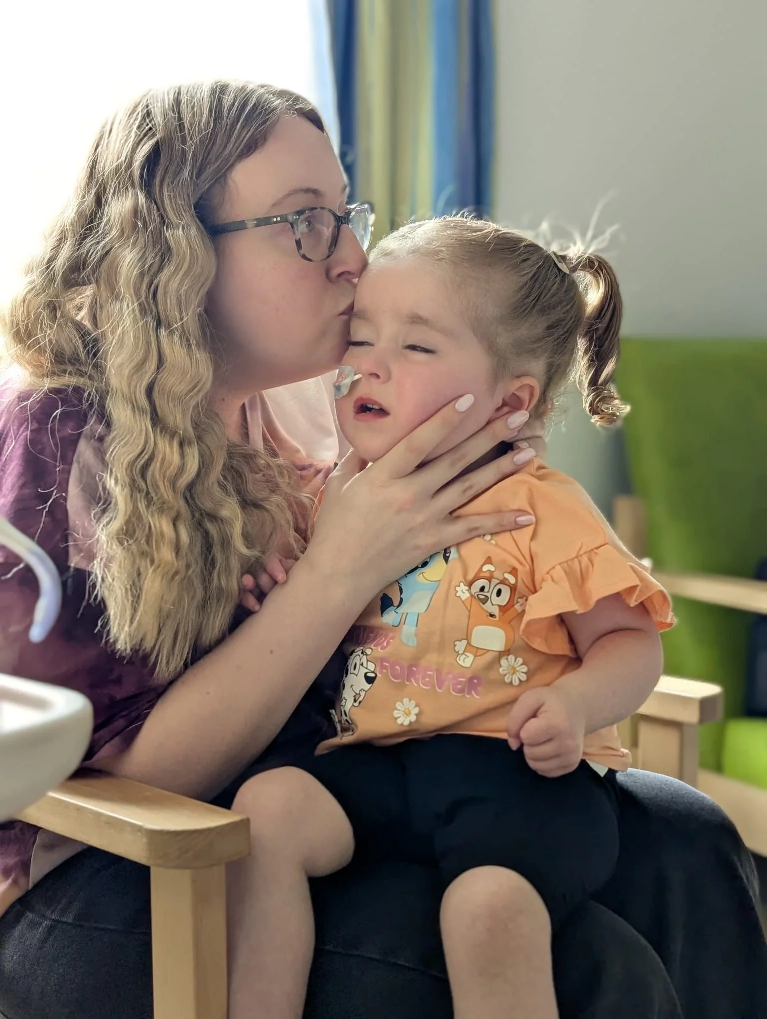 A woman with long curly blonde hair, glasses, and wearing a pink top, is tenderly kissing a young girl on the forehead. The girl has her hair in a ponytail, is wearing an orange shirt with cartoon characters, and appears to be emotionally upset. The woman is holding the girl’s face gently with one hand, and they are sitting close together in a room with light-colored walls and colorful curtains in the background.