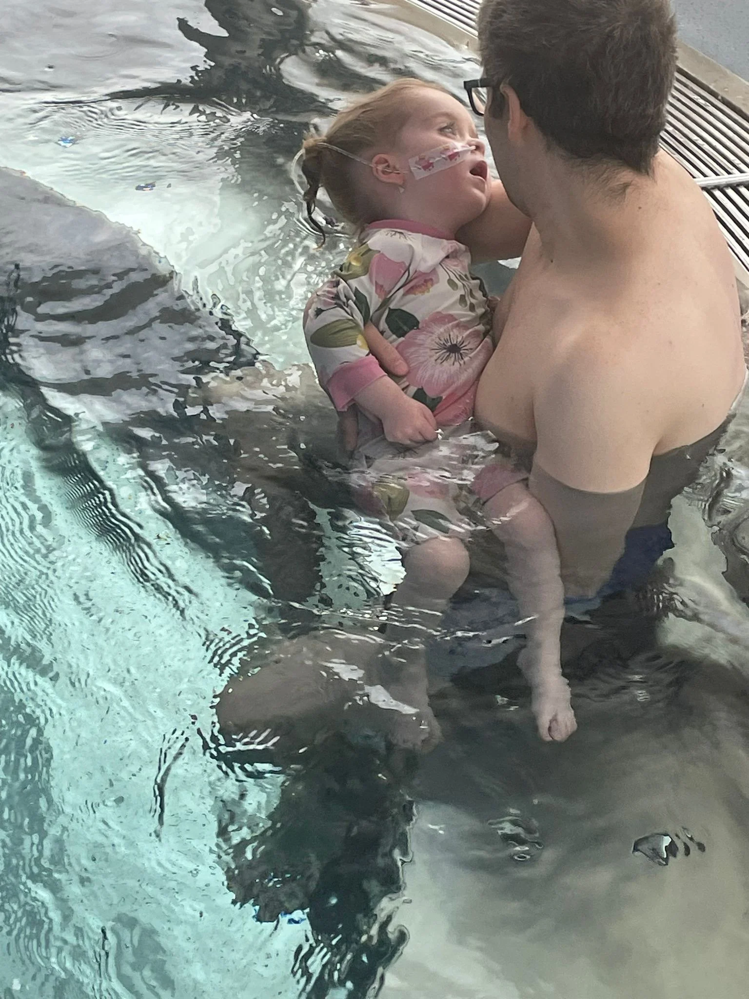A child with a nasal oxygen tube, smiling and looking at a man during a swimming session in a pool. The child is wearing a floral-patterned shirt and appears to be enjoying the moment.