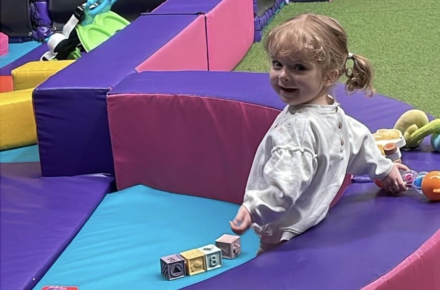 A young girl with blonde curly hair in a ponytail is sitting on a colorful play mat at an indoor playground, smiling at the camera. She is wearing a white sweater and playing with small blocks and toys. The background includes padded walls and foam structures in vibrant colors like purple, pink, blue, yellow, and green.