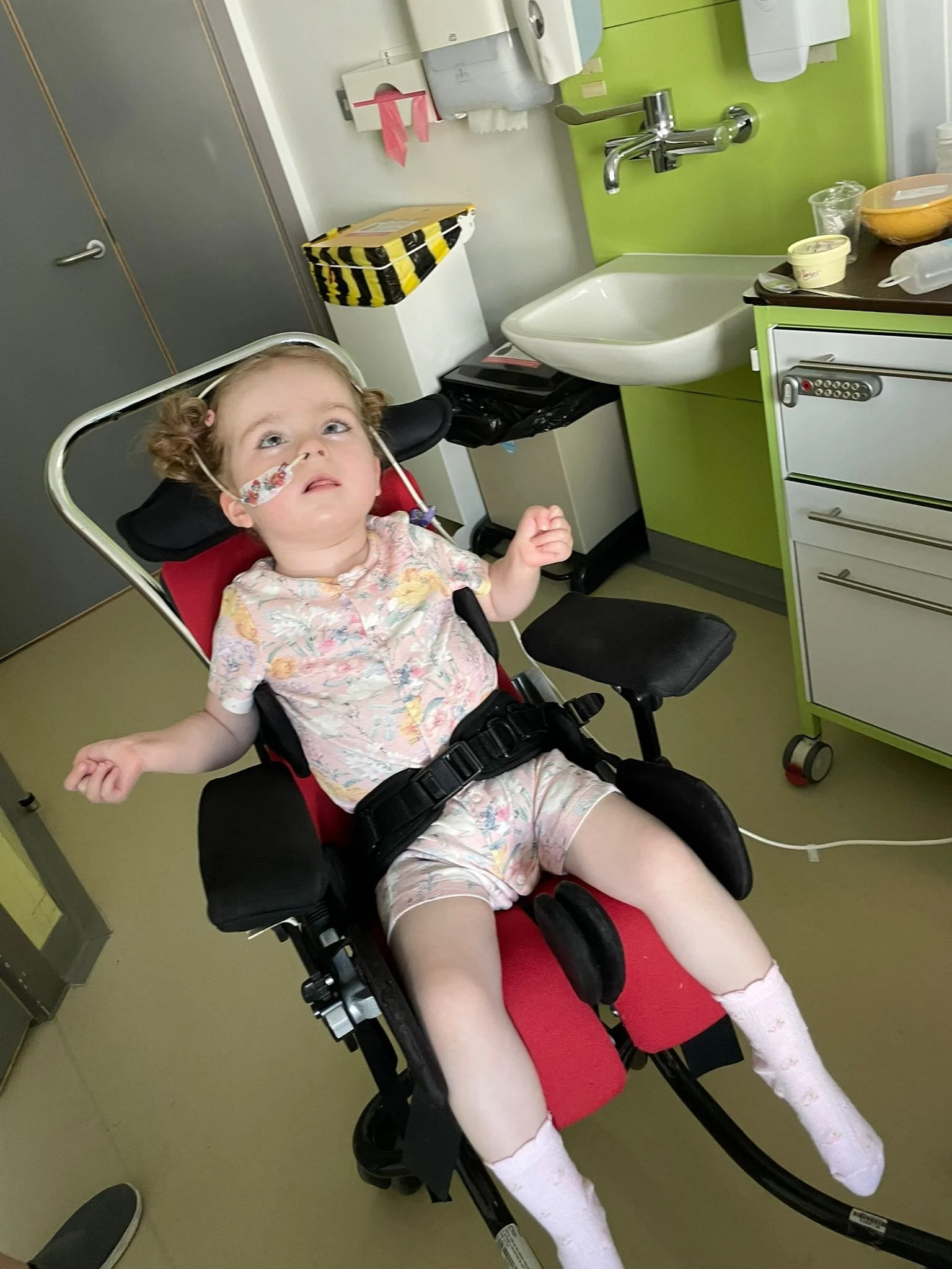 A young girl with curly hair and a nasal cannula lying in a hospital bed in a medical room, with a sink and cabinets in the background.