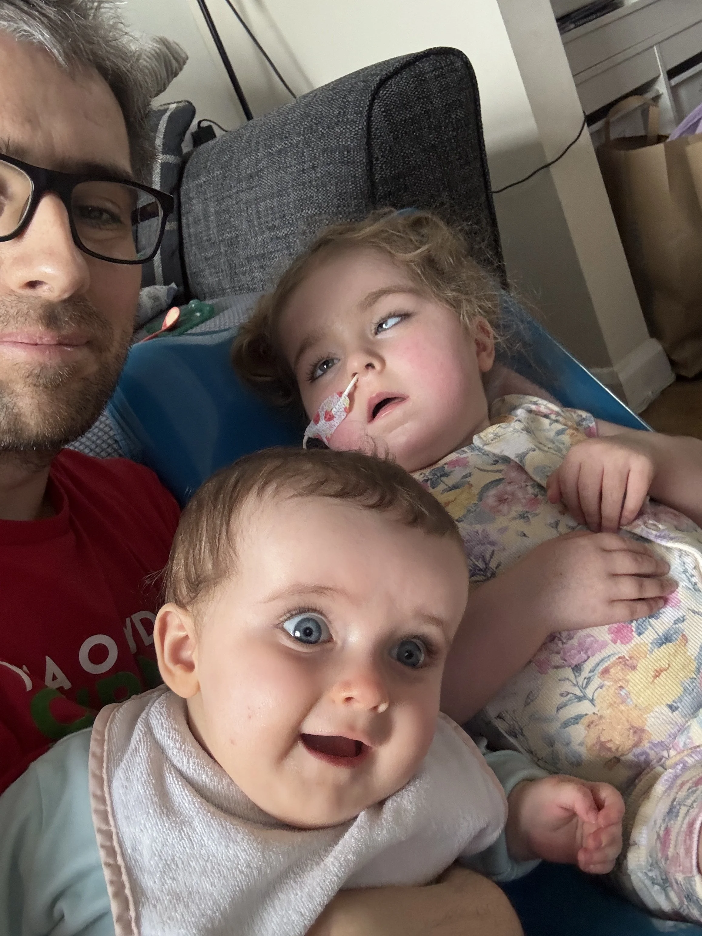 A man with glasses lying next to two young children, one with curly blonde hair and blue eyes, and the other with dark brown hair, both lying on a couch with pillows in the background.