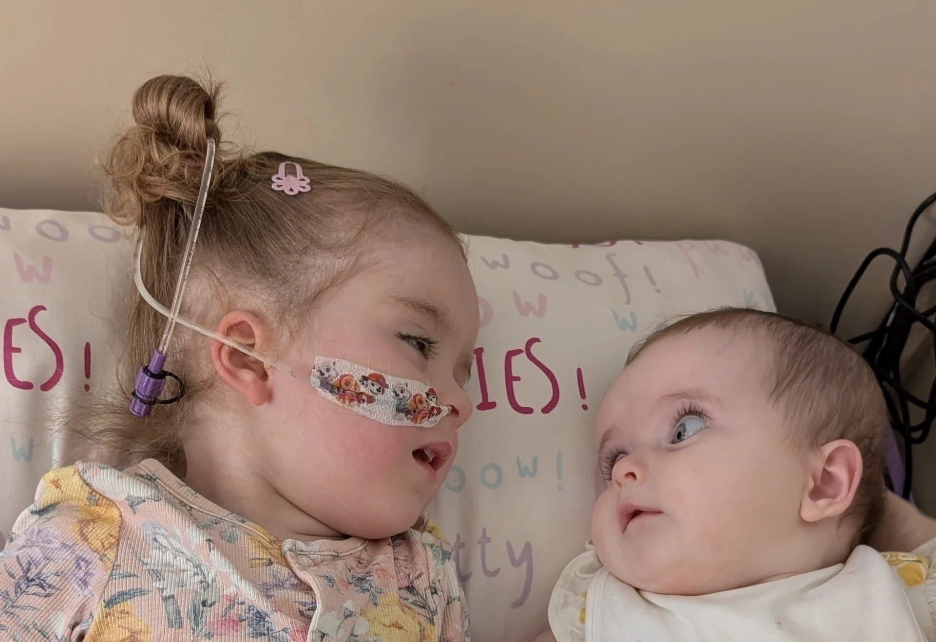 Two young children lying in bed, facing each other and looking into each other's eyes. One girl is wearing a face mask with cartoon characters and has a pink hair clip. The other child, a boy, is looking up at her. The background shows a wall and a bedspread with colorful letters.
