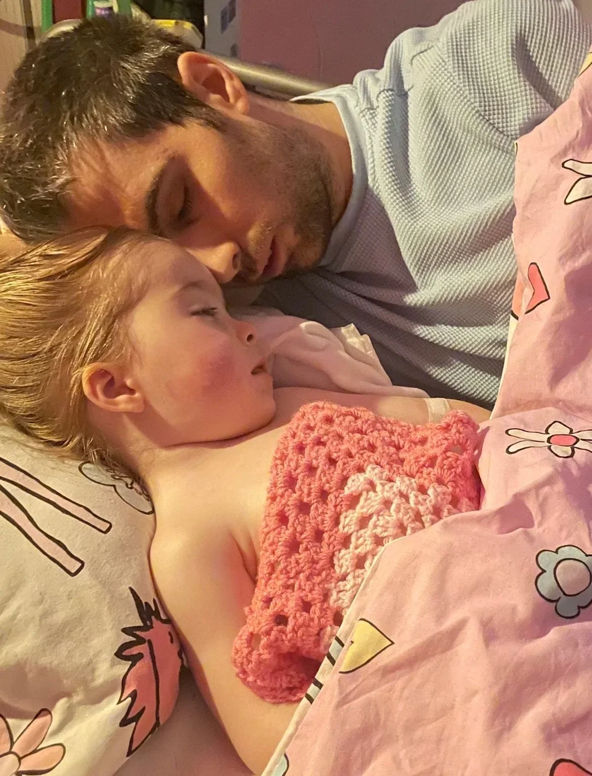A young girl with blonde hair sleeping peacefully in bed next to a man, possibly her father, who is also sleeping with his head resting on the pillow close to hers. They are covered with pink bedsheets decorated with cartoon flowers.