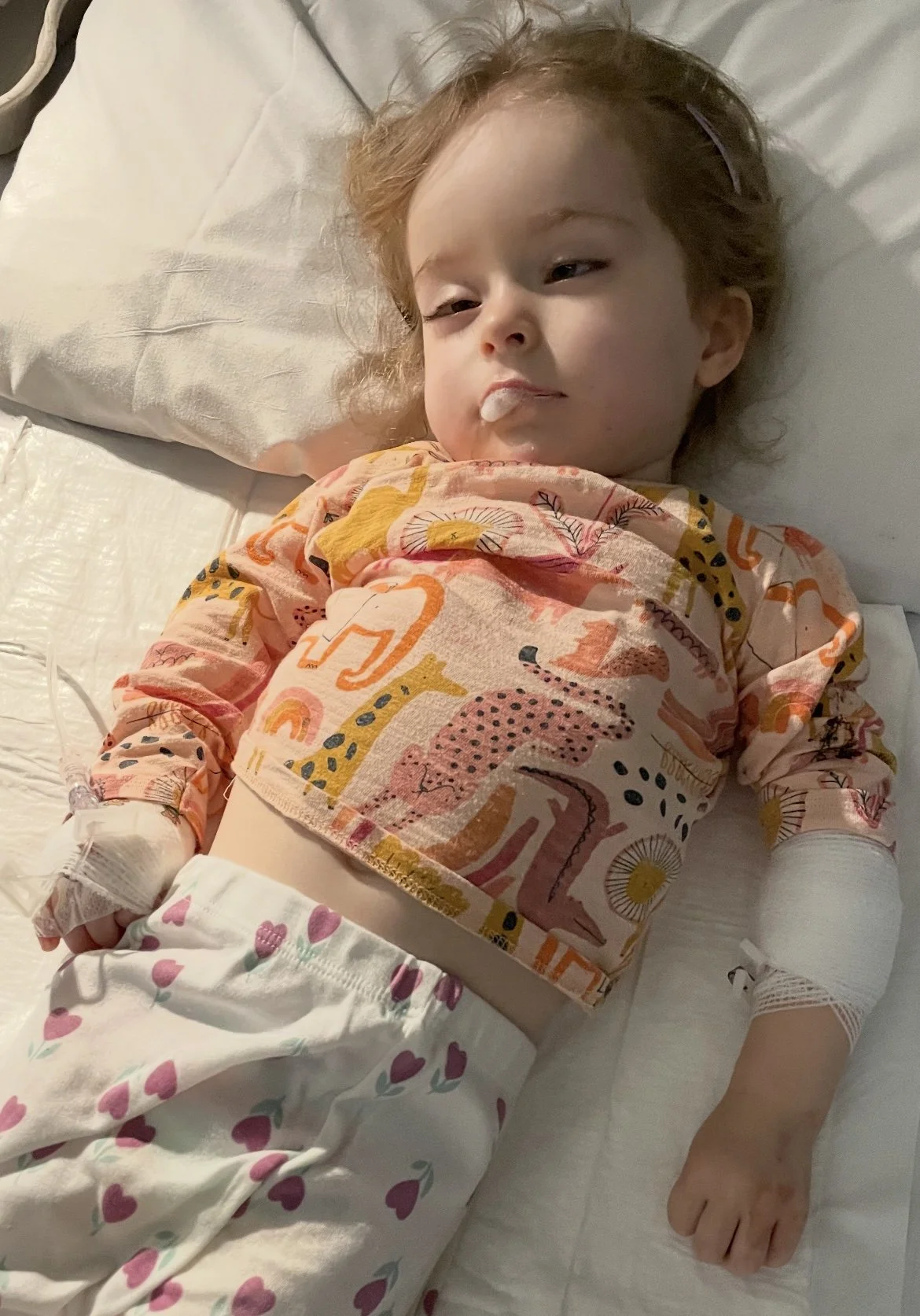 A young child lying in a bed, with IV in their left hand, wearing a colorful shirt with abstract animal patterns and hearts on pants, looking tired or unwell.