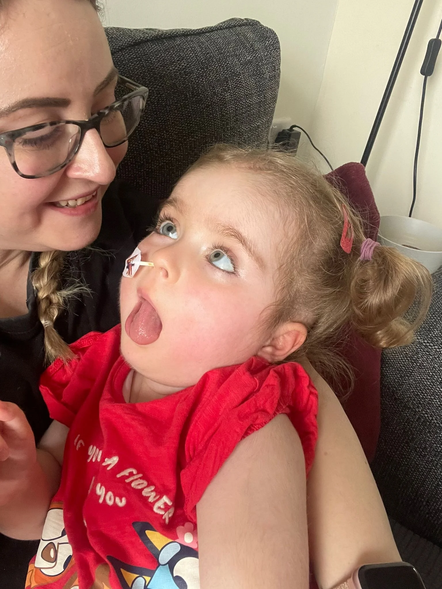 A woman with glasses and braided hair smiles at a young girl with curly hair and blue eyes. The girl is sticking out her tongue and looking up at the woman. They are sitting on a couch with a dark-colored cushion. The girl is wearing a red shirt with a colorful graphic and text.