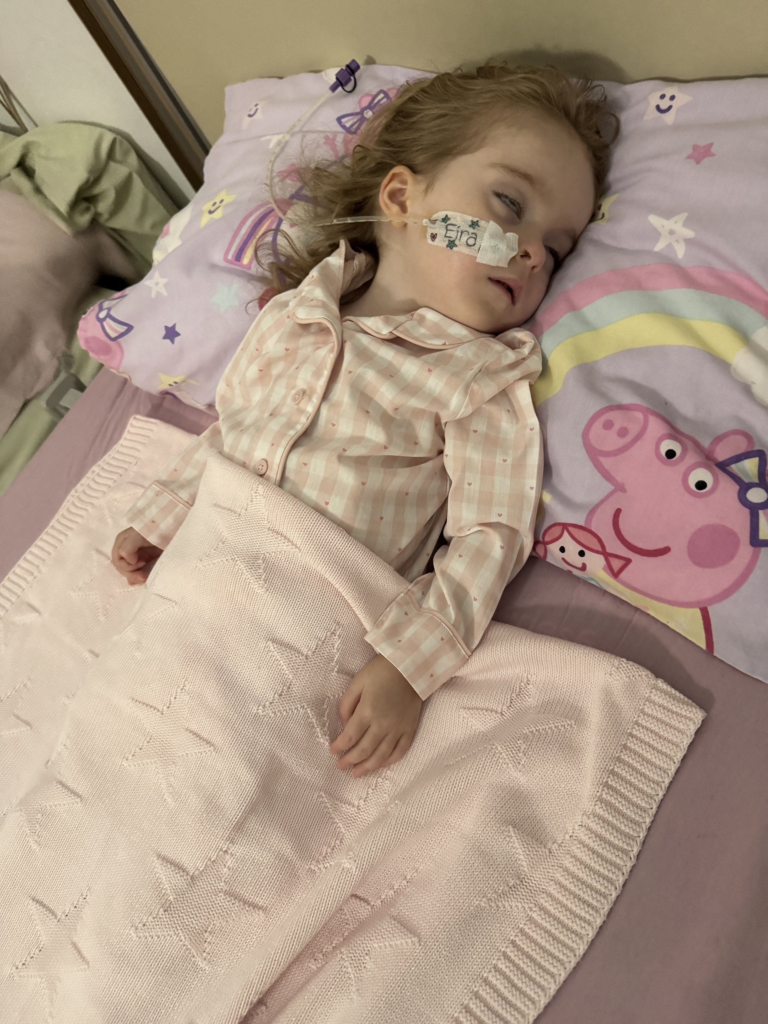 A young girl with red hair lying in a hospital bed with a nasal feeding tube and a name tag that says 'Eira', wearing a pink plaid pajama top. The bed has pink and purple sheets with colorful cartoon characters and stars.