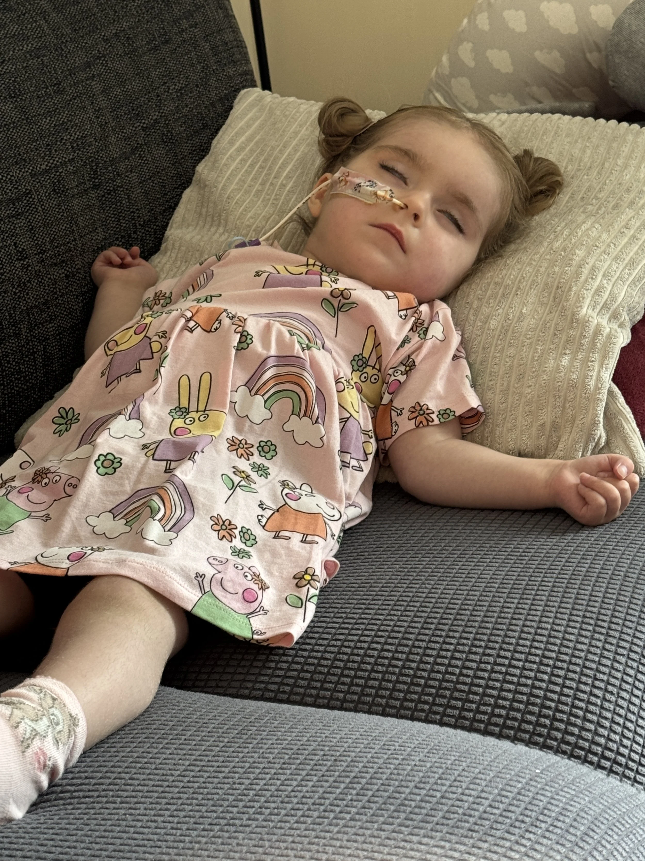 A young girl with light hair styled in two buns is sleeping on a couch, wearing a pink dress with colorful cartoon bunny and rainbow patterns. She has a nasal cannula for oxygen and is lying on a beige textured pillow.