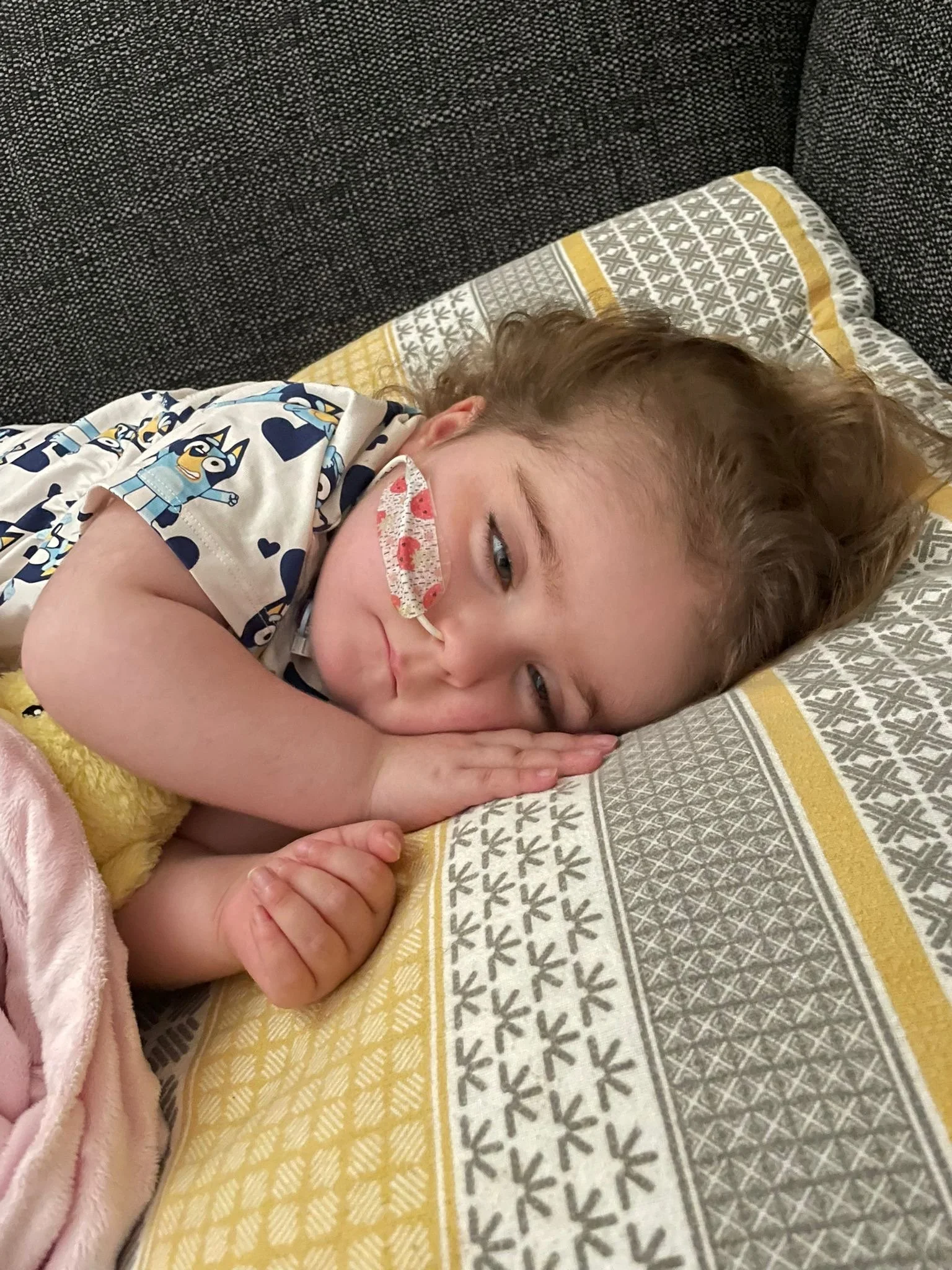 A child with light brown hair, wearing a face mask and a pajama with cartoon characters, is lying on a patterned pillow and blanket on a dark gray couch.