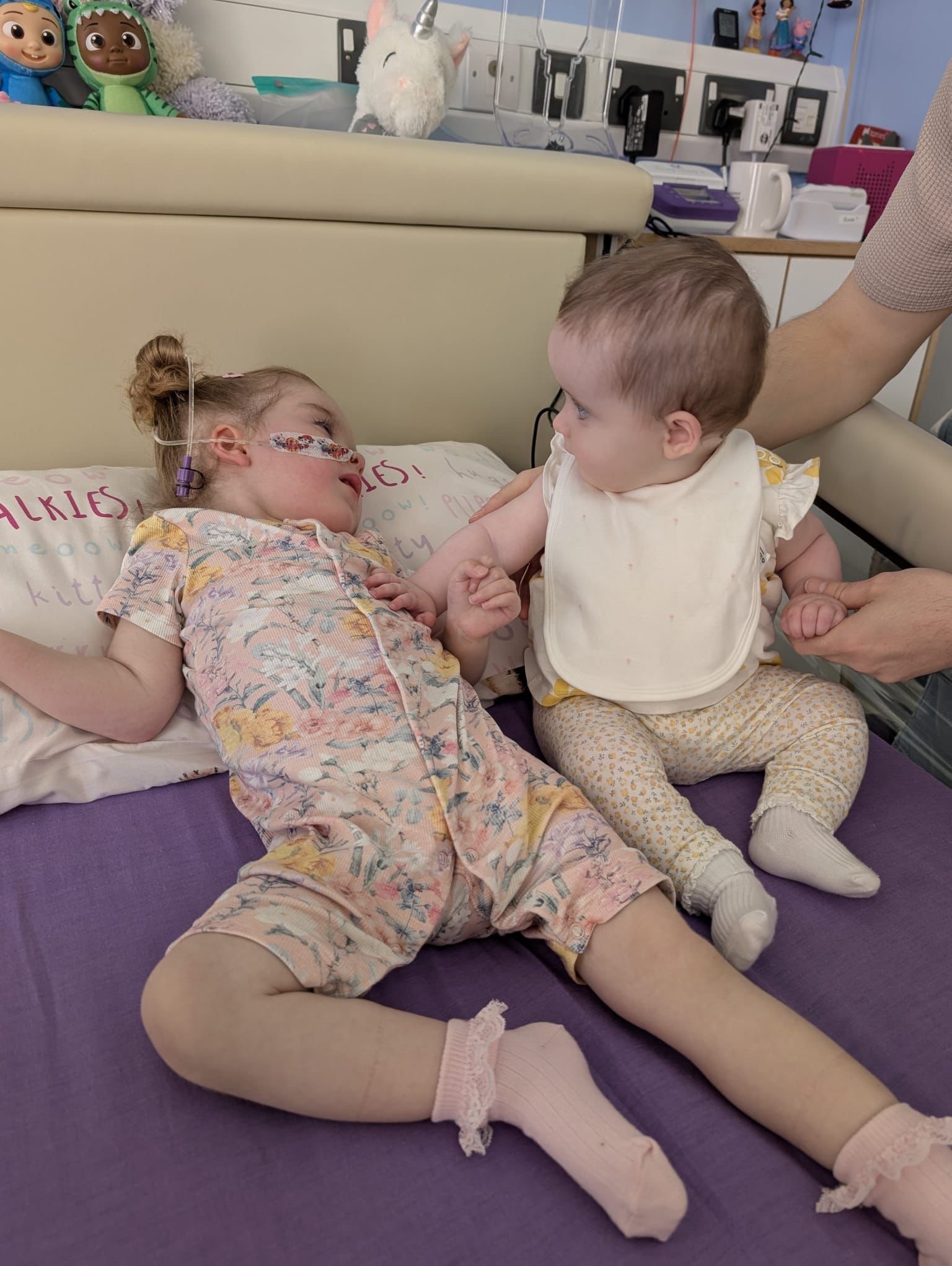 A young girl lying in a hospital bed with a nasal cannula, playing with a small child who is sitting on her legs and touching her face. The girl has her hair in a bun and wears pajamas with a floral pattern. The child has short hair, a bib, and patterned pants.
