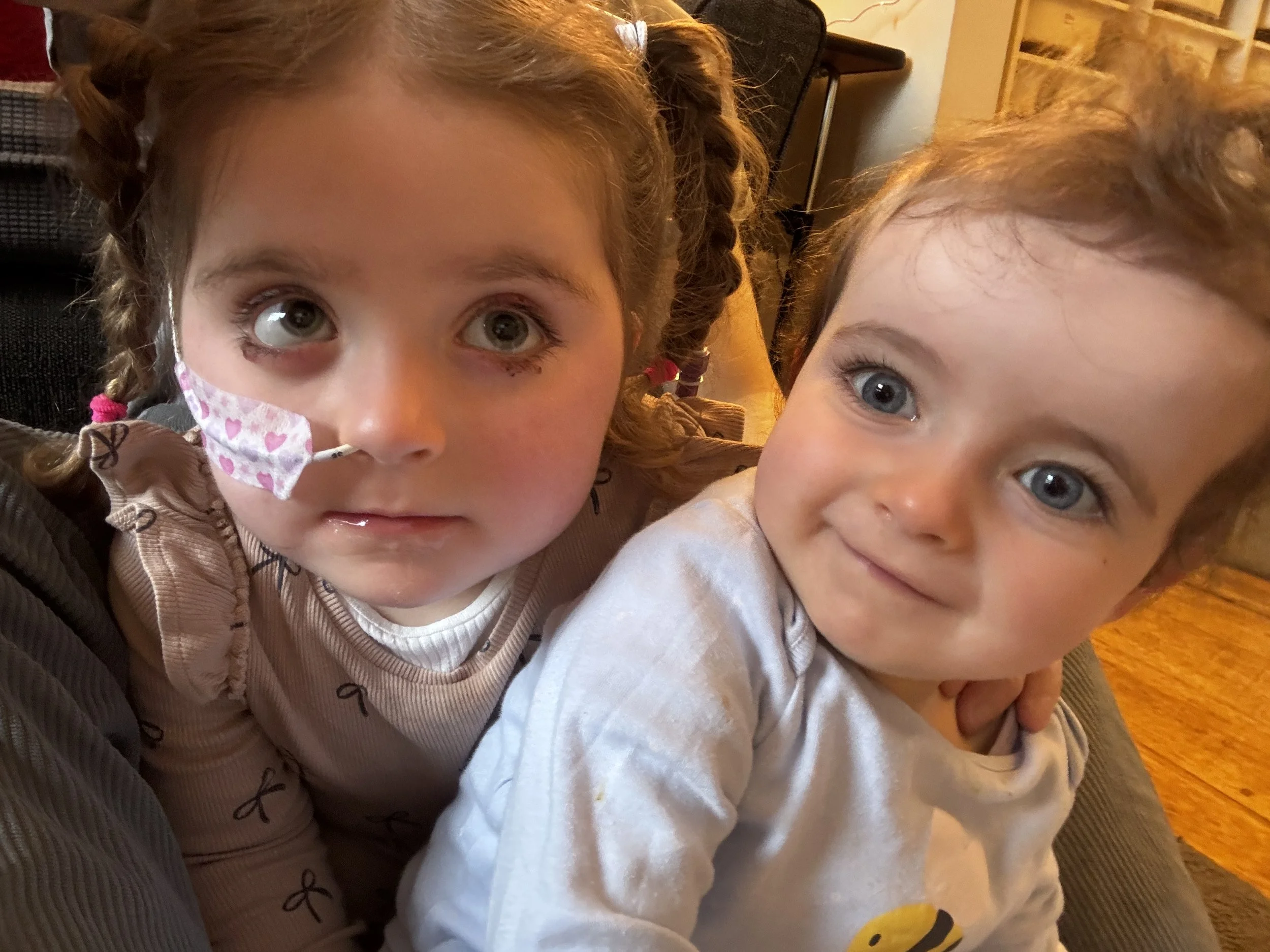 Two young girls with curly hair and blue eyes close together, smiling and looking up. The girl on the left is wearing a face mask with pink hearts and a beige jacket. The girl on the right is wearing a light-colored shirt. They are indoors, with a piece of furniture and a yellow and orange patterned carpet visible in the background.