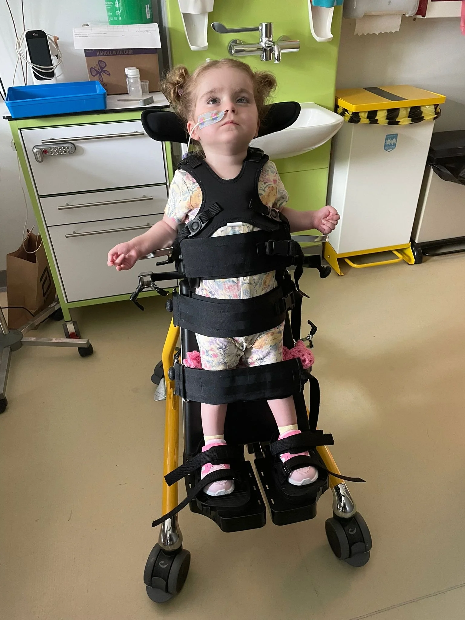 Young girl in a wheelchair with a nasal oxygen tube, standing in a medical setting, with medical supplies and furniture in the background.