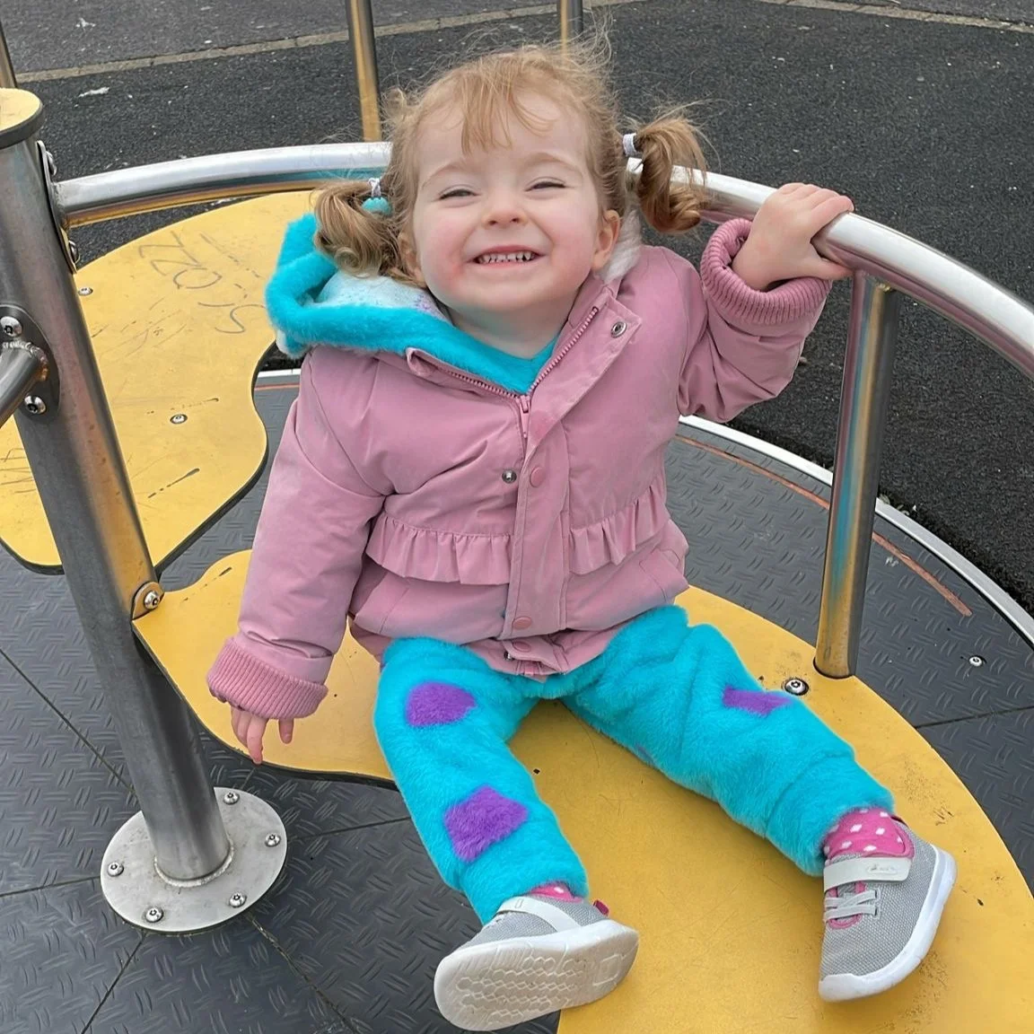 A young girl with pigtails smiling and playing on a carousel at a playground, wearing a pink jacket, blue fleece pants with purple hearts, and sneakers.