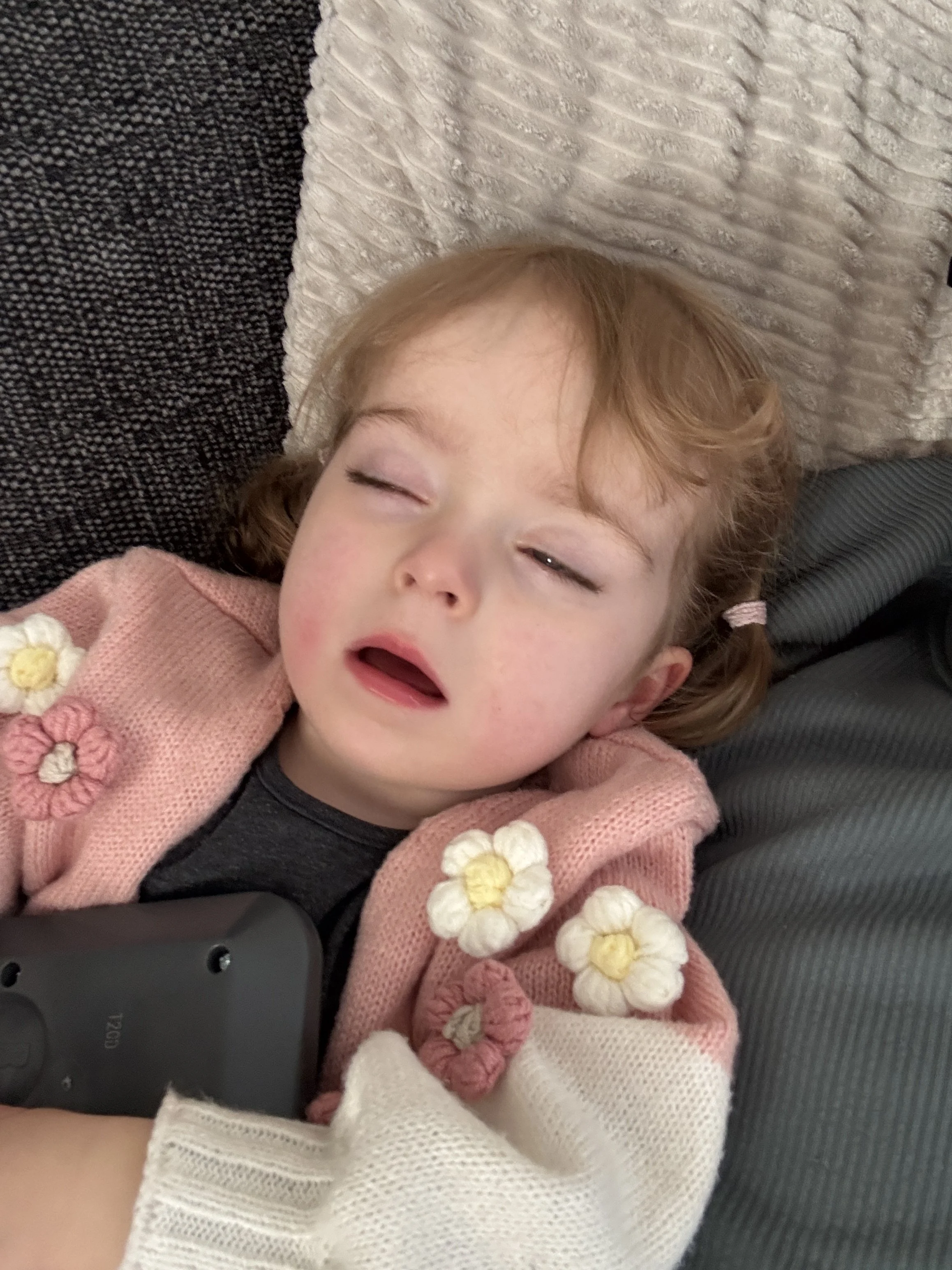A young girl with curly red hair sleeping on a bed, wearing a pink cardigan with flower embellishments, with a black remote control partially visible at the bottom of the image.