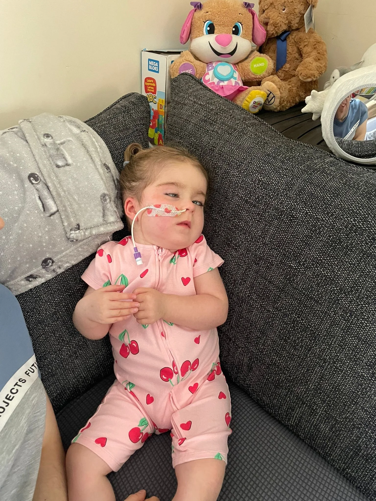 A young girl with a medical tube in her nose, lying on a black textured couch. She is wearing a pink outfit with cherries and green leaves and appears to be resting with her eyes partially open. There are plush toys, including a large character with a pink bow and a teddy bear, positioned behind her.