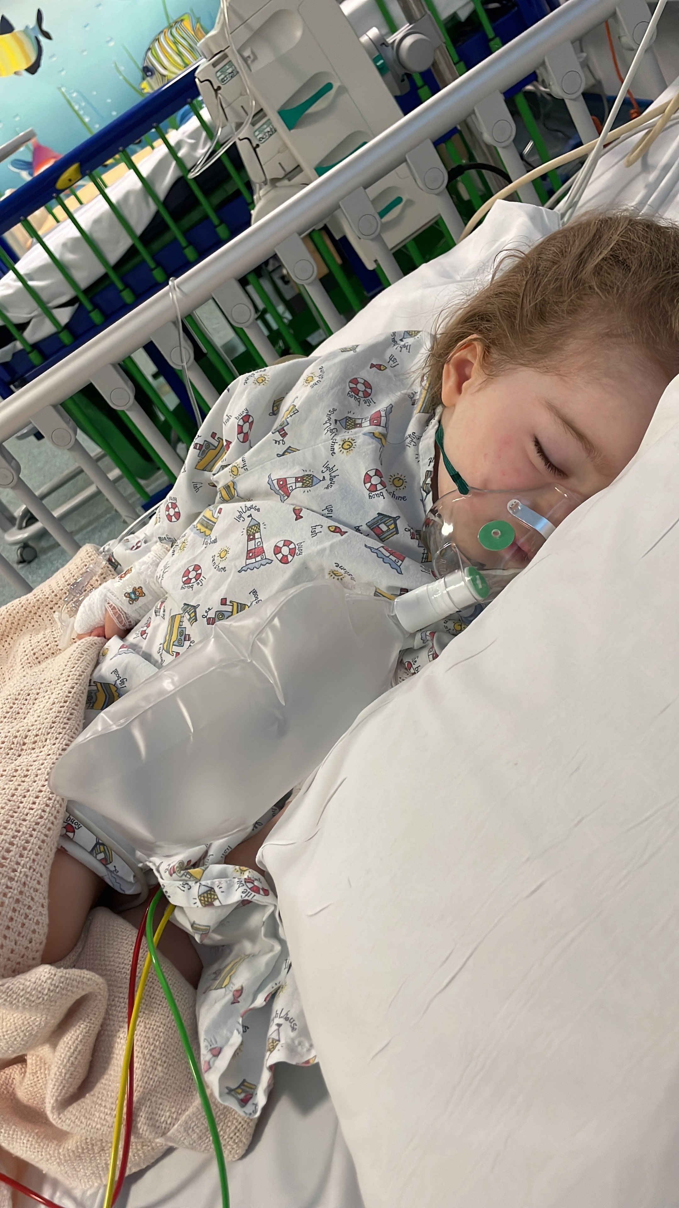 Child lying on hospital bed with oxygen mask, hospital gown with nautical theme, and IV in hand, in a pediatric hospital room.
