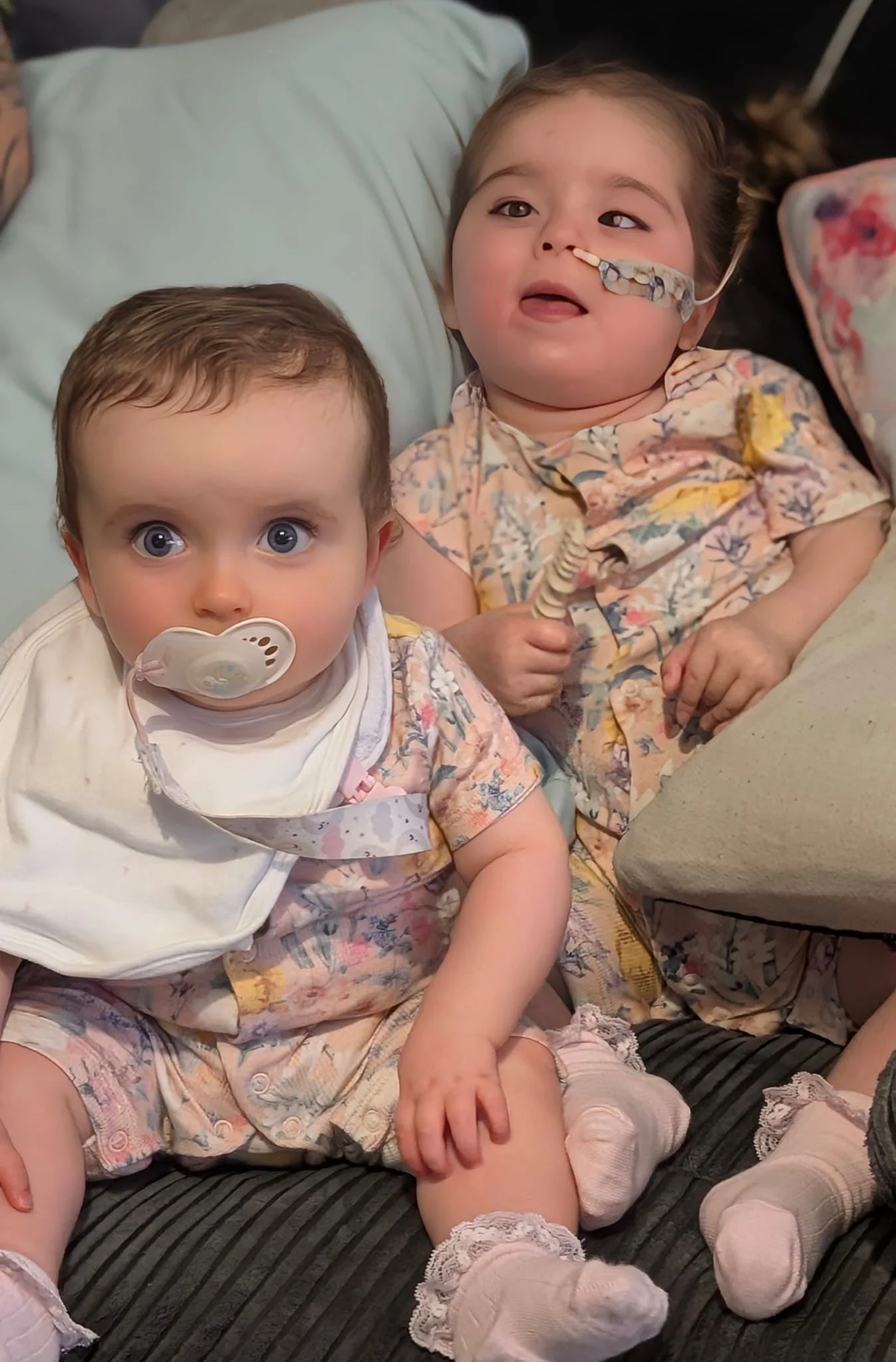 Two young children, a girl with a nasal tube and a pacifier, and a boy with blue eyes and no visible medical devices, sitting on a striped black sofa in floral pajamas. The girl is smiling and the boy is looking directly at the camera.