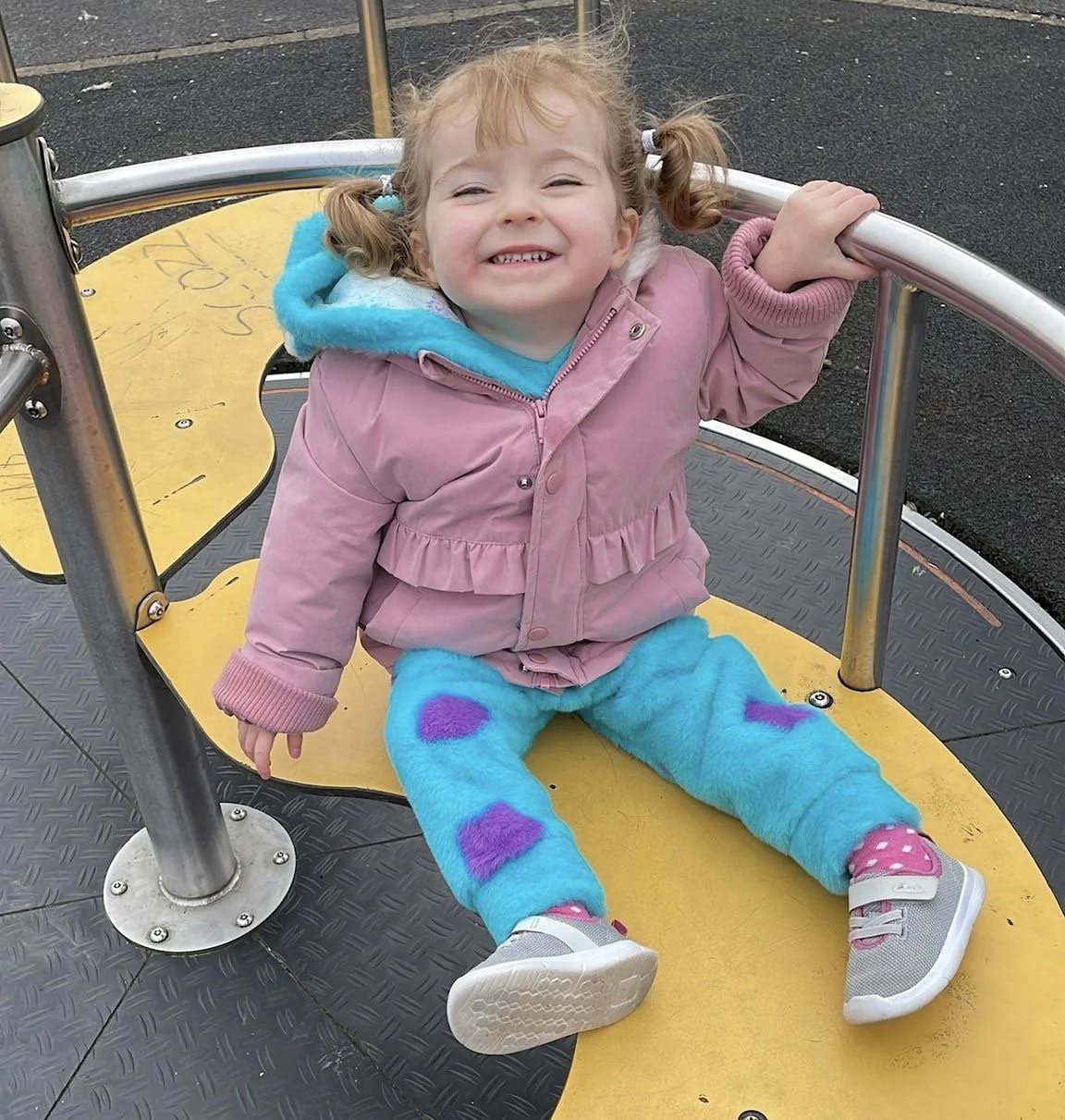 A young girl with curly hair in pigtails smiling while sitting on a yellow playground ride with metal railings at a park.