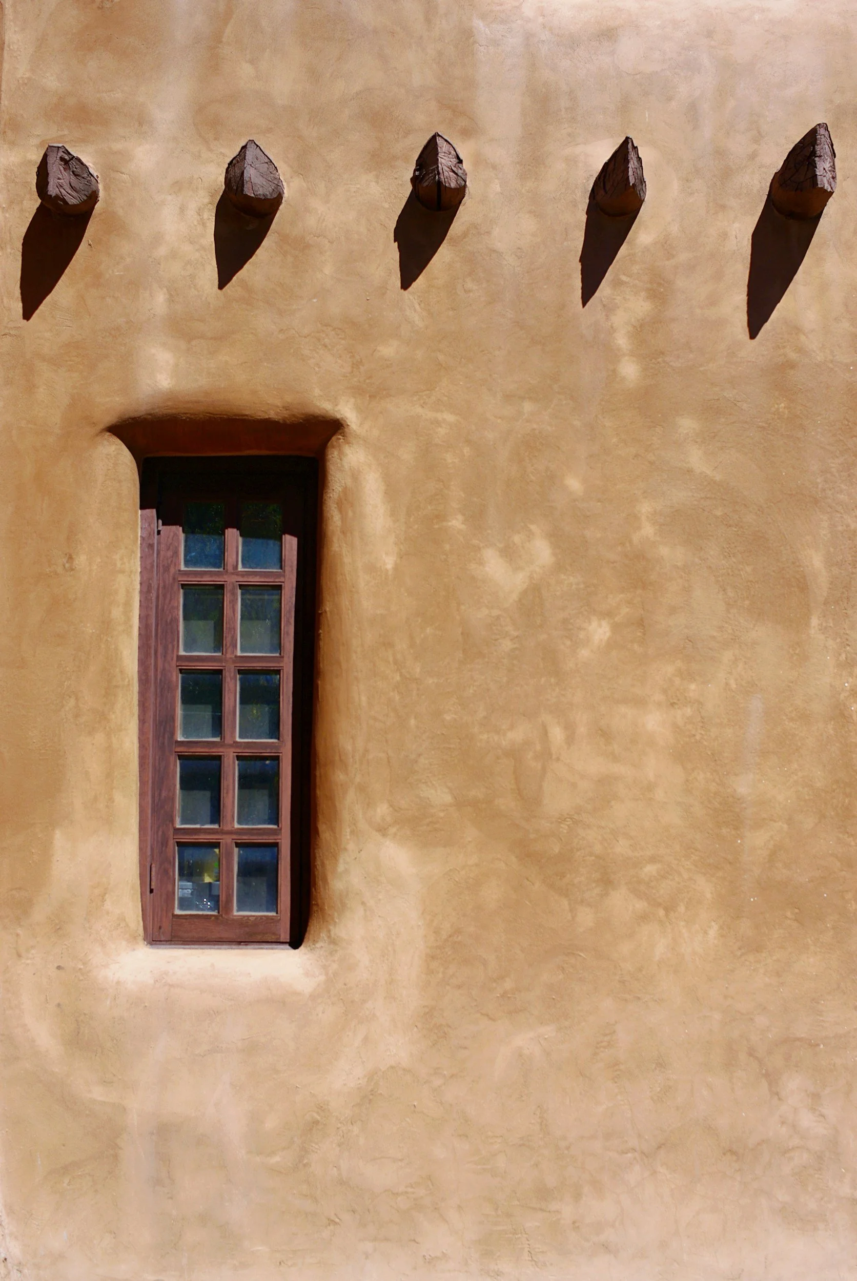 A brown stucco building wall with a single rectangular wooden-framed window and four wooden pegs protruding from the wall above.