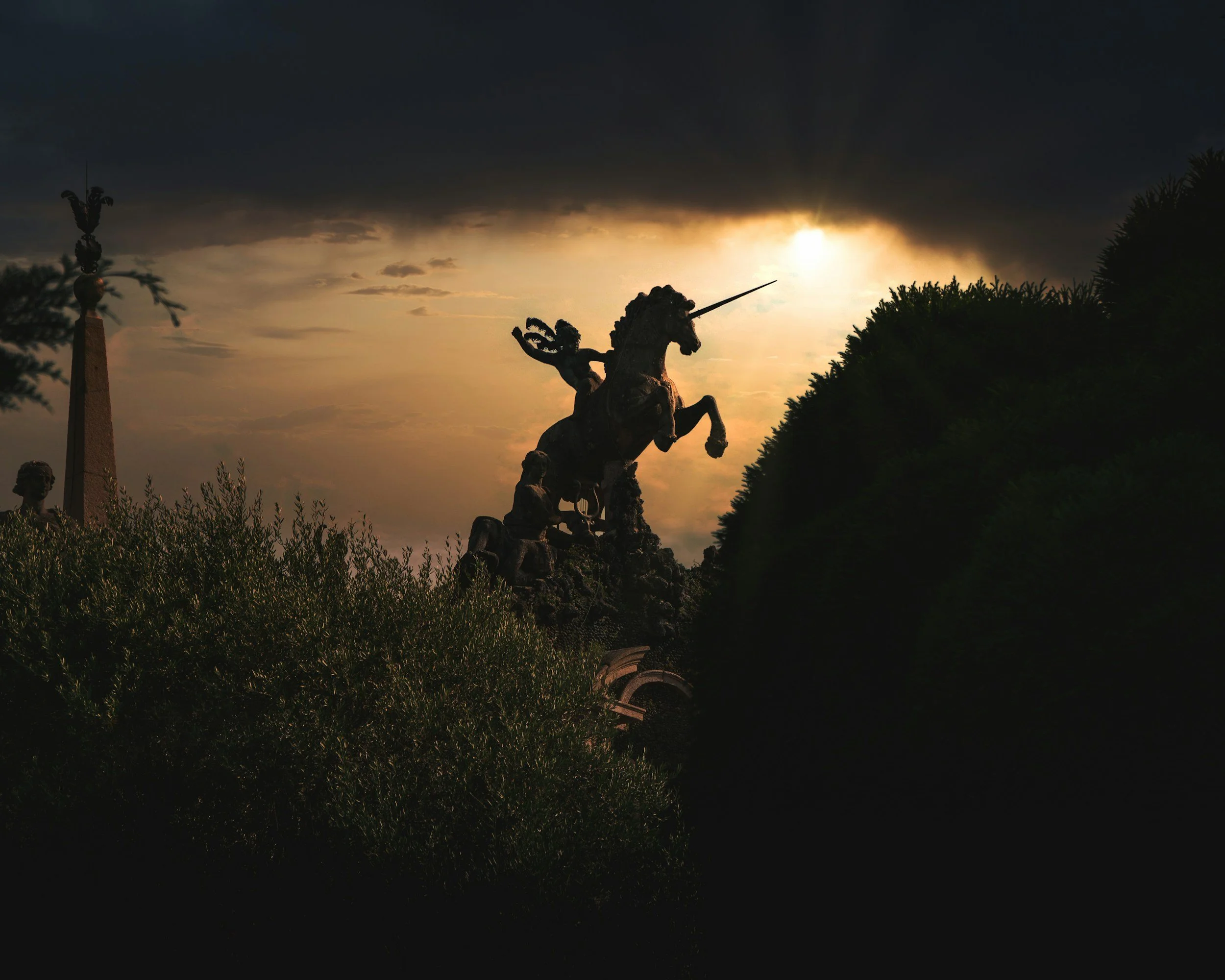 A bronze statue of a man riding a horse and holding a spear, set against a dramatic sunset sky with dark clouds, greenery in the foreground, and additional statues or monument structures visible at the edges.