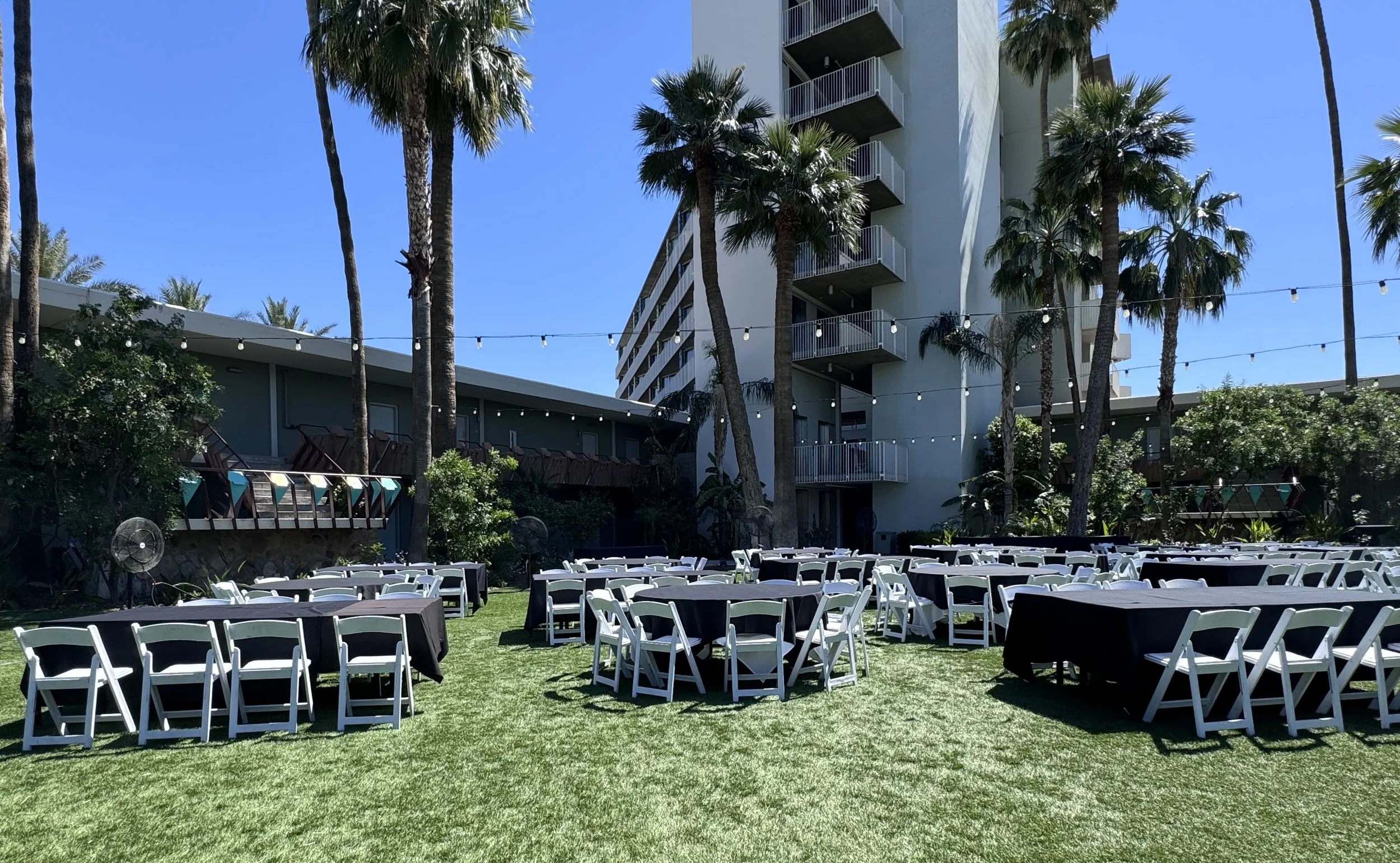 Outdoor event setup with rows of chairs and tables on green grass, palm trees, string lights, and a modern building in the background.