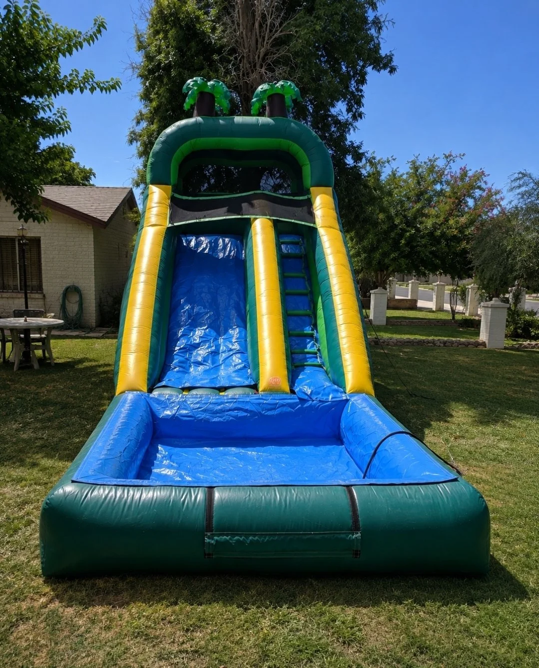 Colorful inflatable water slide with a blue sliding surface, yellow sides, and green decorative elements on top, set up on a grassy yard on a sunny day.