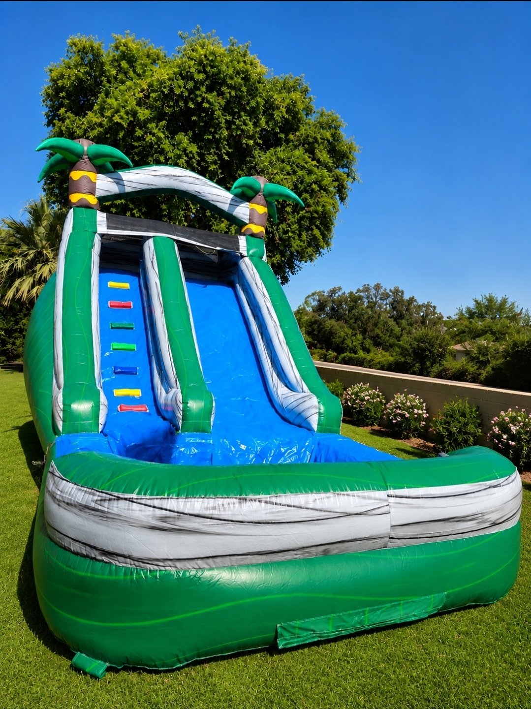 Inflatable water slide featuring a tropical theme with palm trees at the top, set outdoors on green grass with trees and bushes in the background, under a clear blue sky.