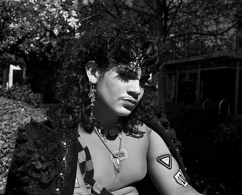Black and white photo of a queer person with curly hair wearing earrings, layered necklaces, and a jacket with badges, sitting outdoors with trees in the background.