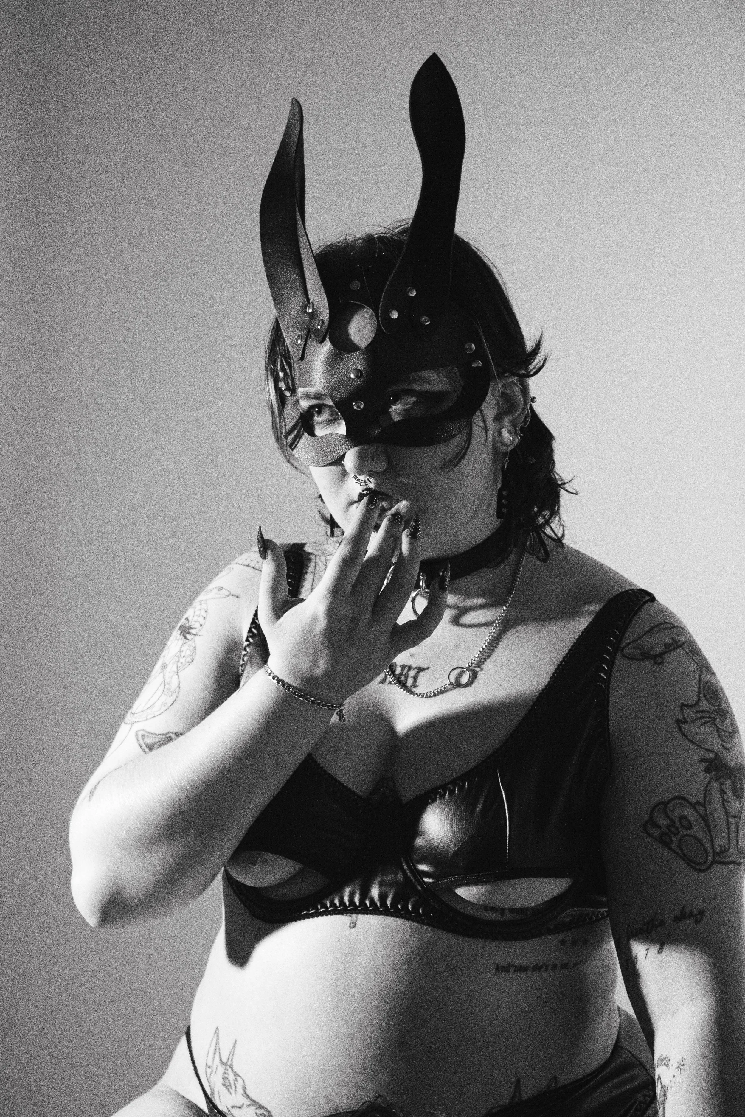 A woman wearing a black mask with bunny ears and a nose, posing with her hand near her face. She has tattoos on her arms and torso, and is dressed in a leather top and jewelry, in a black and white photo.