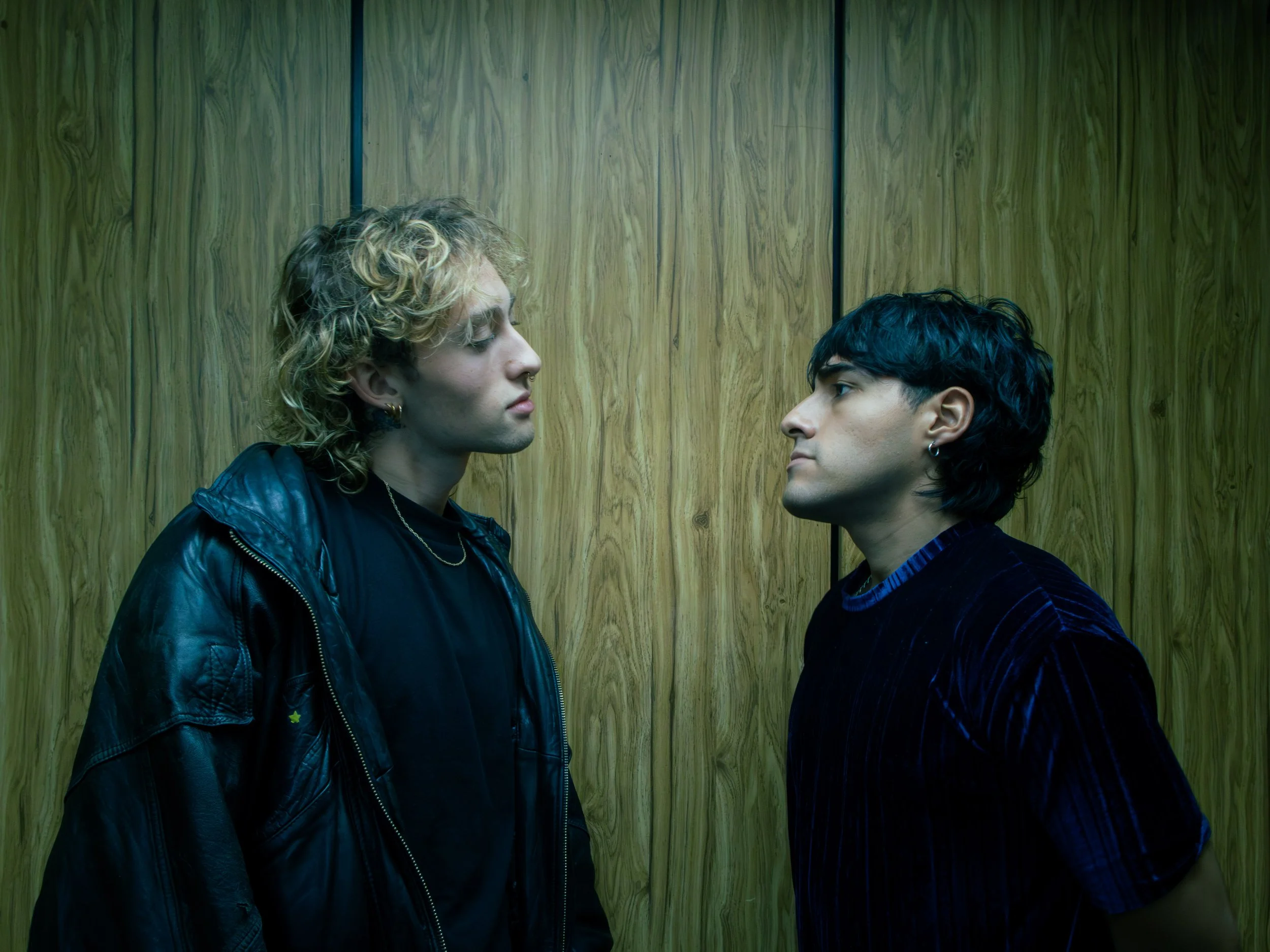 Two men face each other closely inside an elevator with wood-paneled walls.