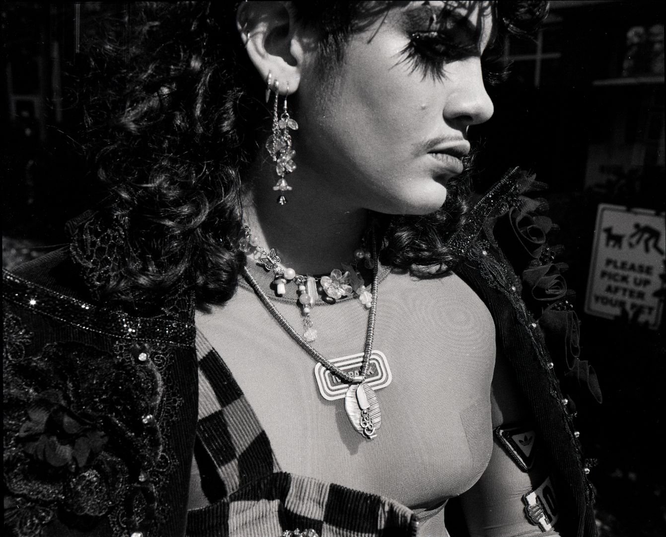 Close-up black and white photo of a queer person with curly hair wearing multiple necklaces and earrings, dressed in a checkered top and decorated jacket, standing in a dark setting.