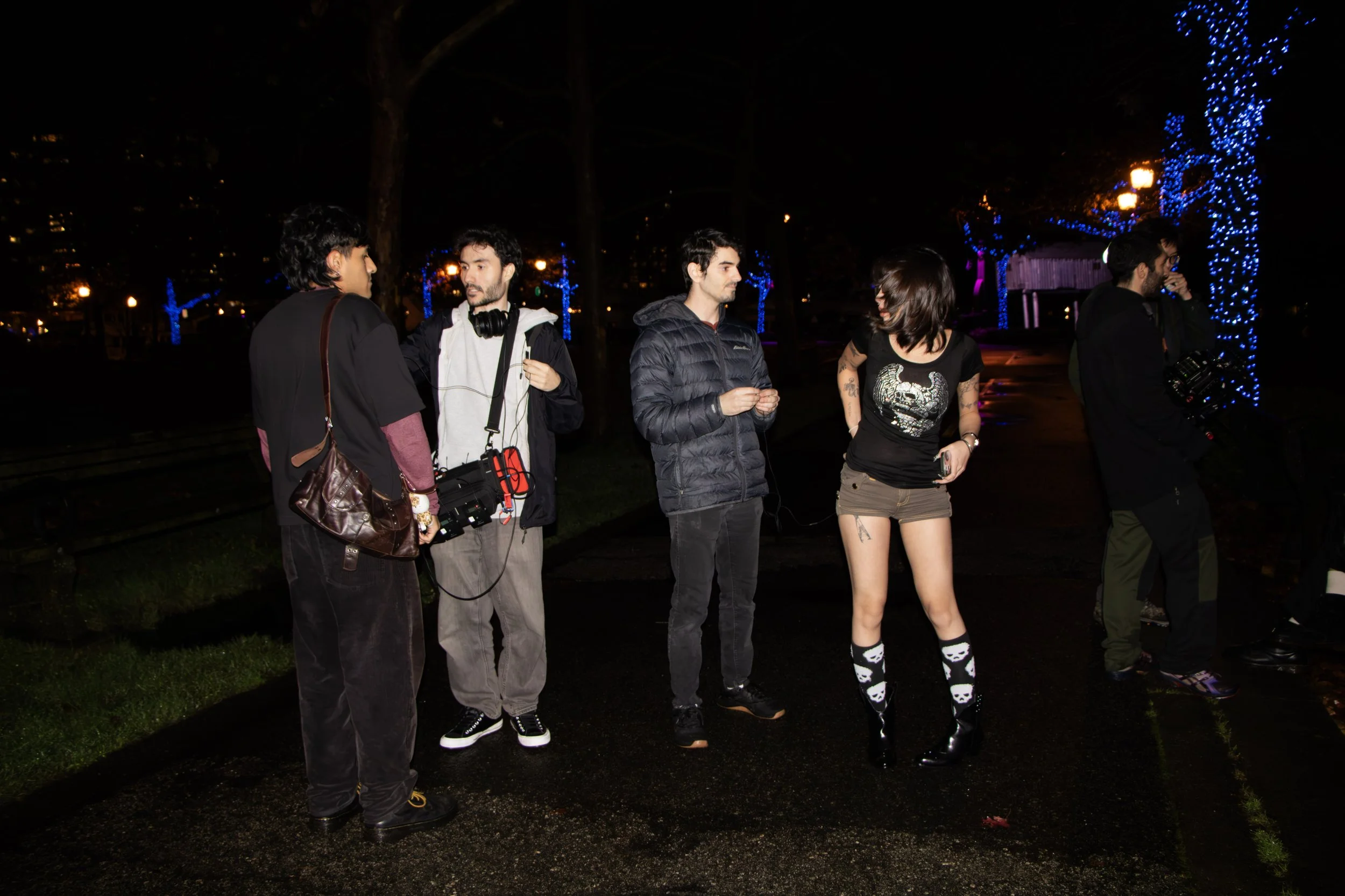 A group of five young adults standing outdoors at night, surrounded by trees decorated with blue lights. Three men and two women are engaged in conversation, with one woman wearing a black skull-printed shirt and black shoes with skull socks.