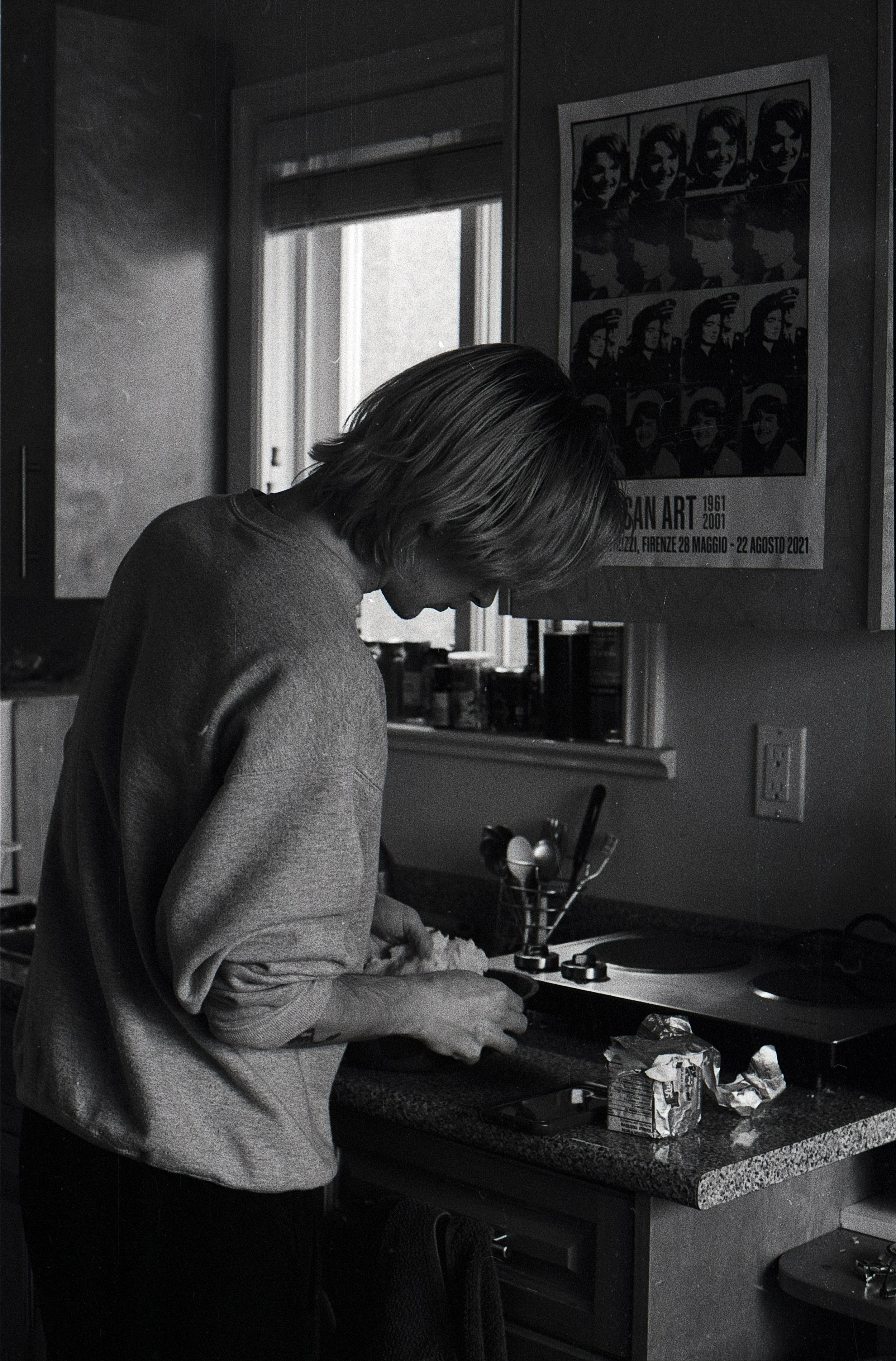 A person with shoulder-length hair wearing a sweatshirt, working with their hands at a kitchen counter, with a bowl and crumpled butter packaging on the counter. There is a window and a poster on the shelf.