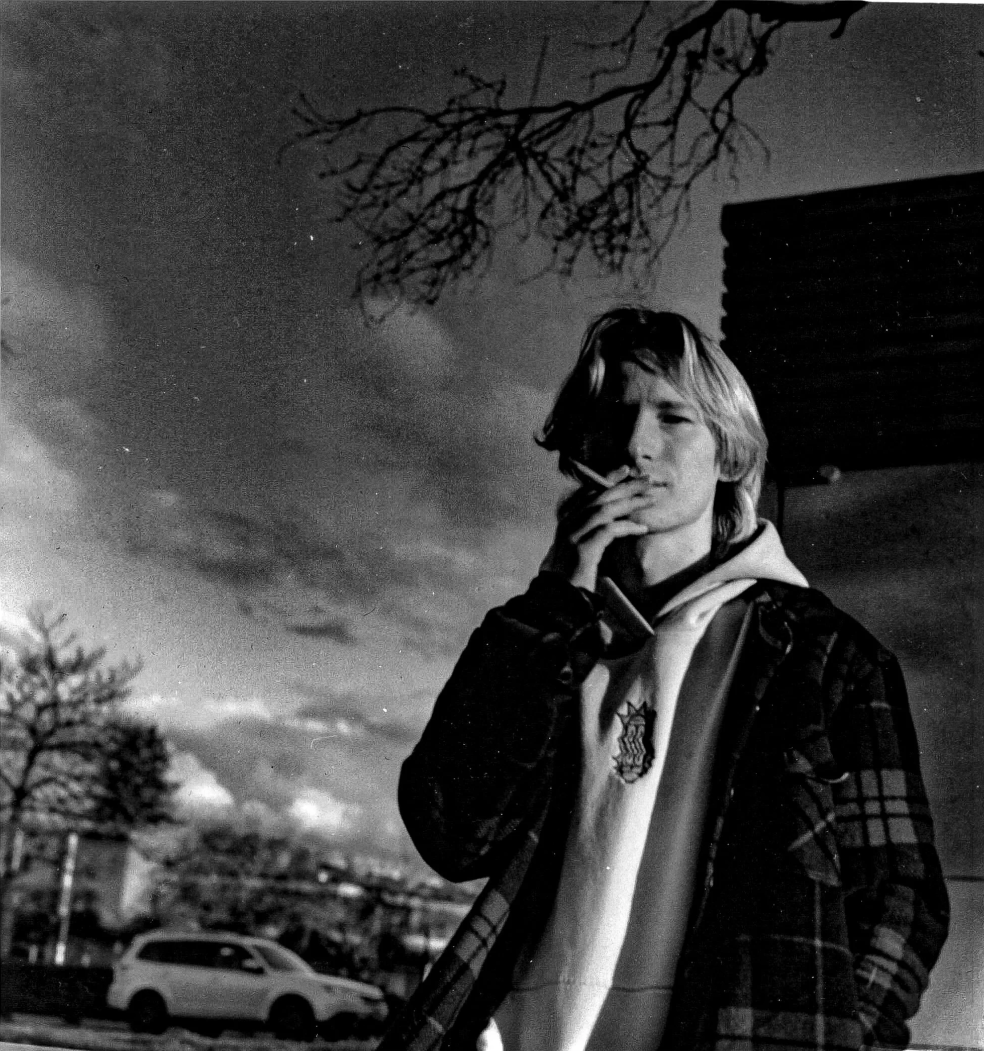 A person standing outdoors while smoking. There's a tree and a building in the background, and a parked car nearby.