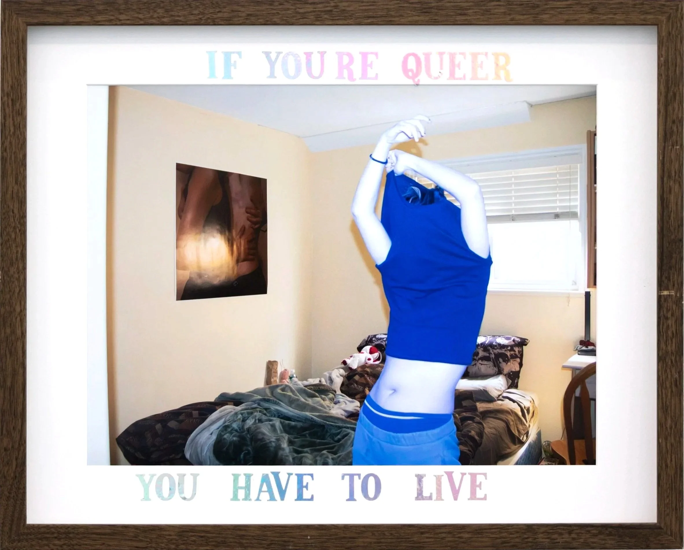 Framed wall art with the text 'If you're queer you have to live' at the top and 'You have to live' at the bottom, overlooking a bedroom with unmade bed and window.