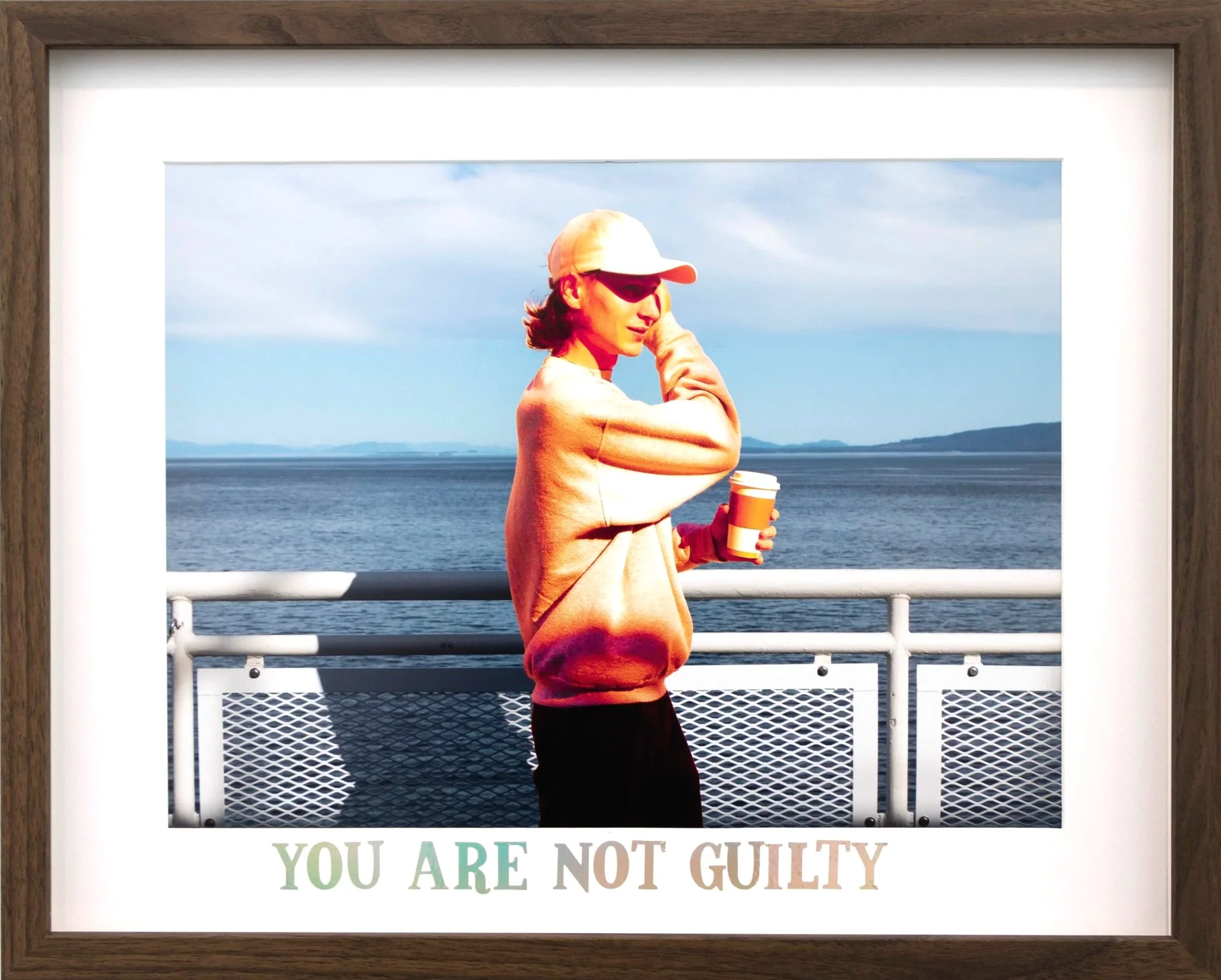 A woman standing by a railing near the water, wearing a beige hat, sunglasses, and a beige sweater, holding a coffee cup, with the ocean and sky in the background.