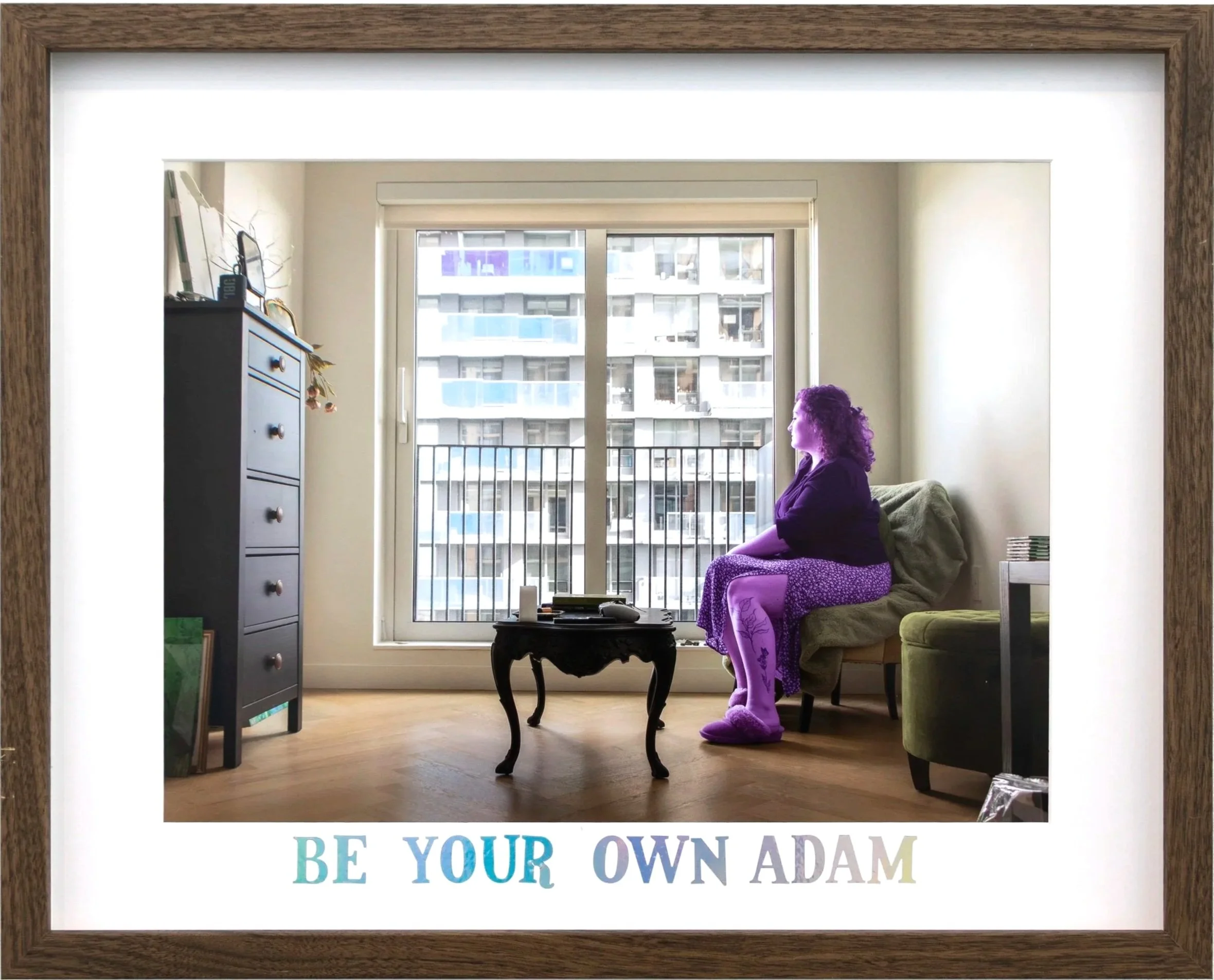 Framed photo showing a woman with purple curly hair sitting on a green couch in a living room by a large window. There is a blue chest of drawers and a small black table in the room. The window reveals a cityscape with multiple apartment buildings. P
