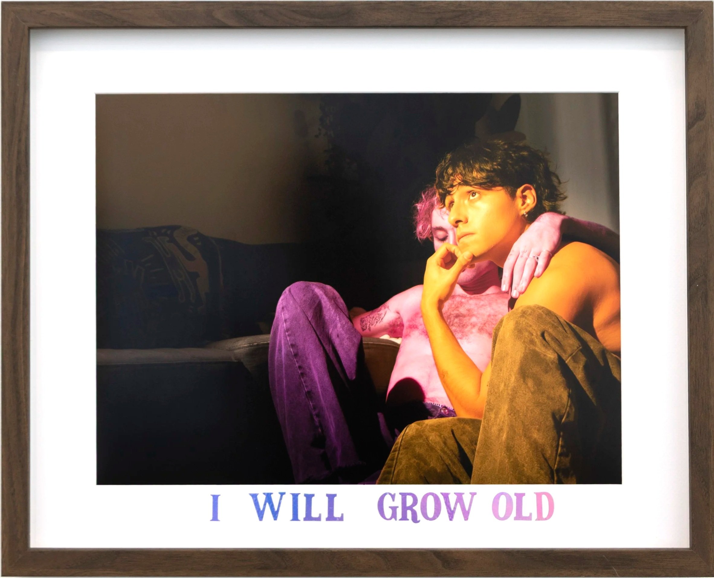 A framed photograph of two young people sitting close together, one with pink hair and the other with dark curly hair, with a sign below that says 'I WILL GROW OLD'.