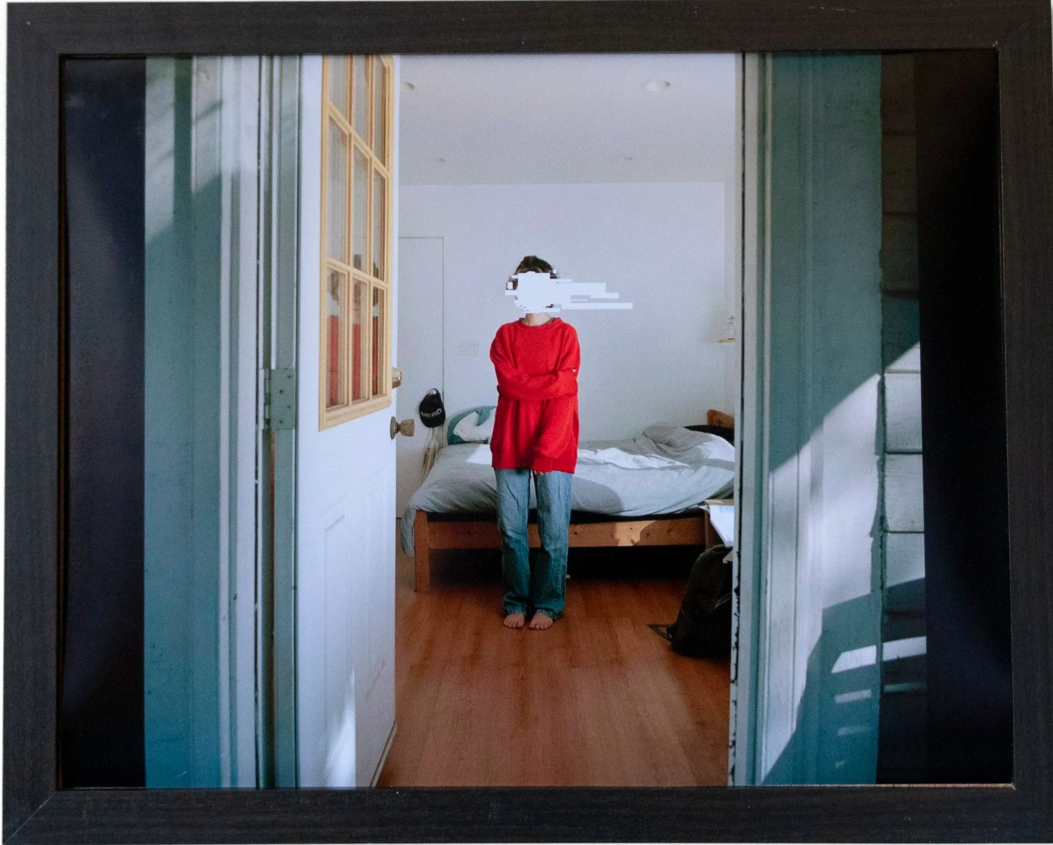 A person wearing a red top and jeans standing barefoot in a bedroom, taking a mirror selfie. The room has a bed, a window, and some personal items visible.