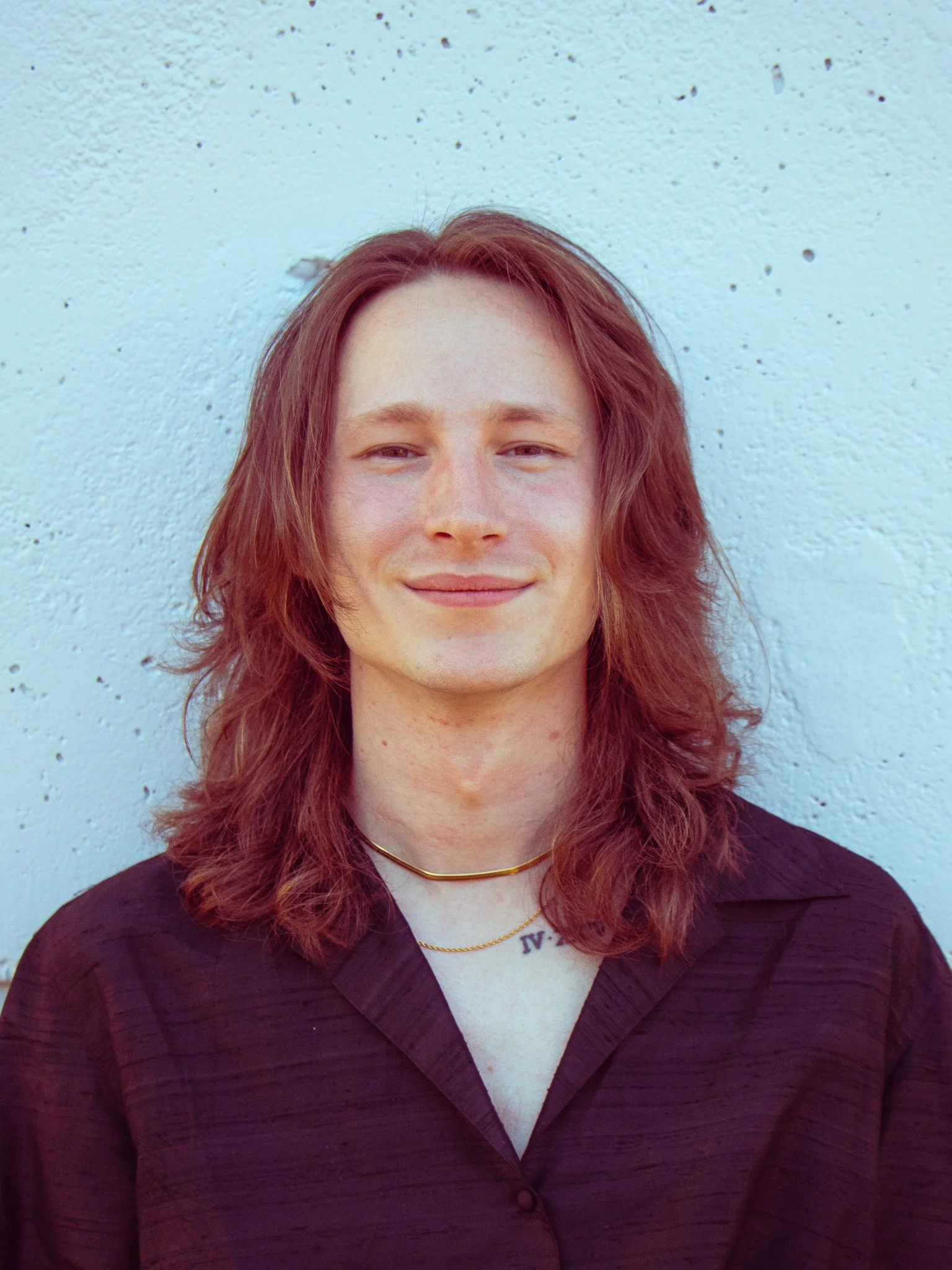A young man with long red hair, wearing a dark shirt, gold necklaces, and a white top underneath, standing against a light blue wall with small black spots, smiling slightly.